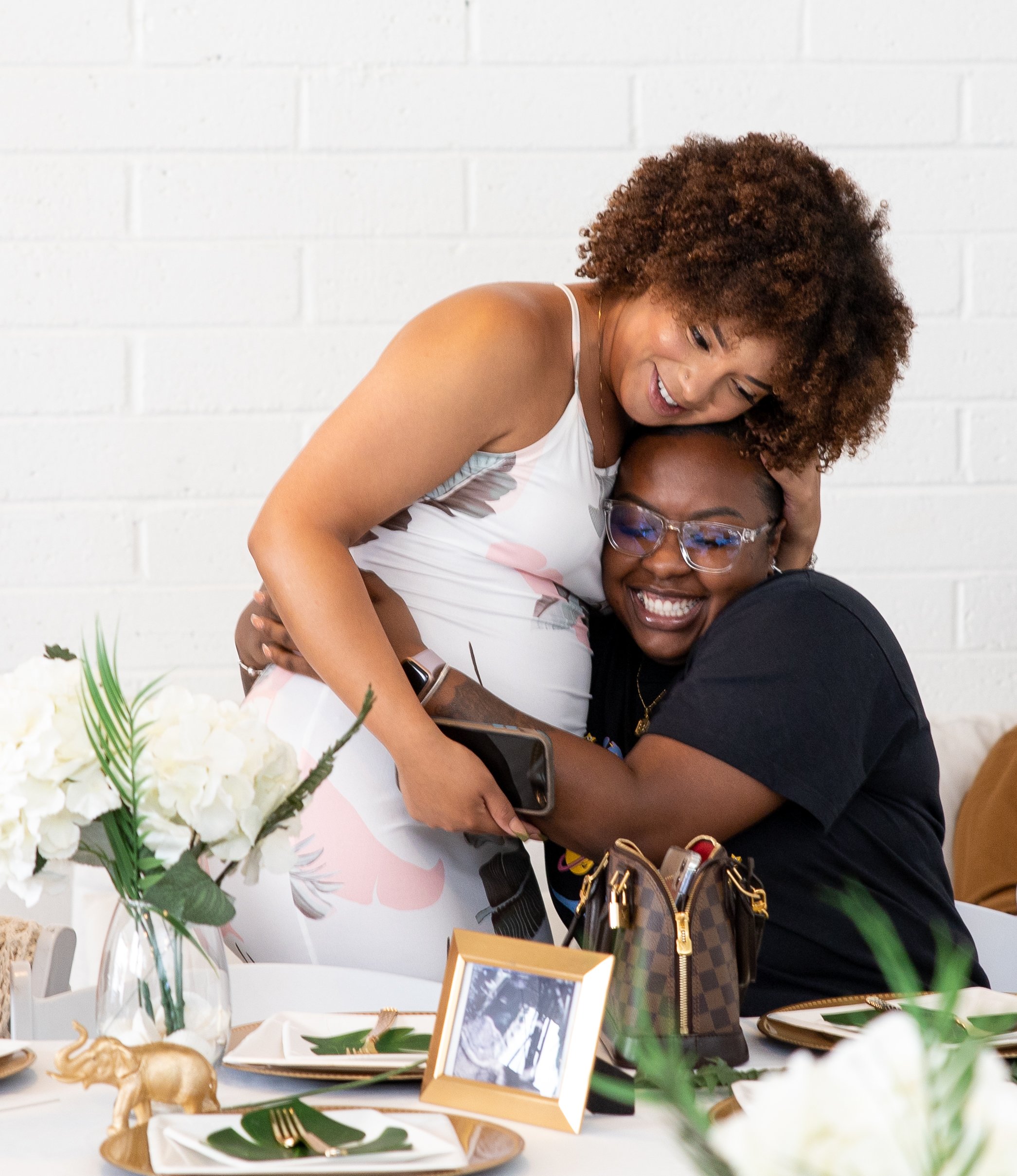 Two women sharing a joyful moment at a celebration, one seated and the other hugging her from above, with a decorated table and floral arrangements in the background.