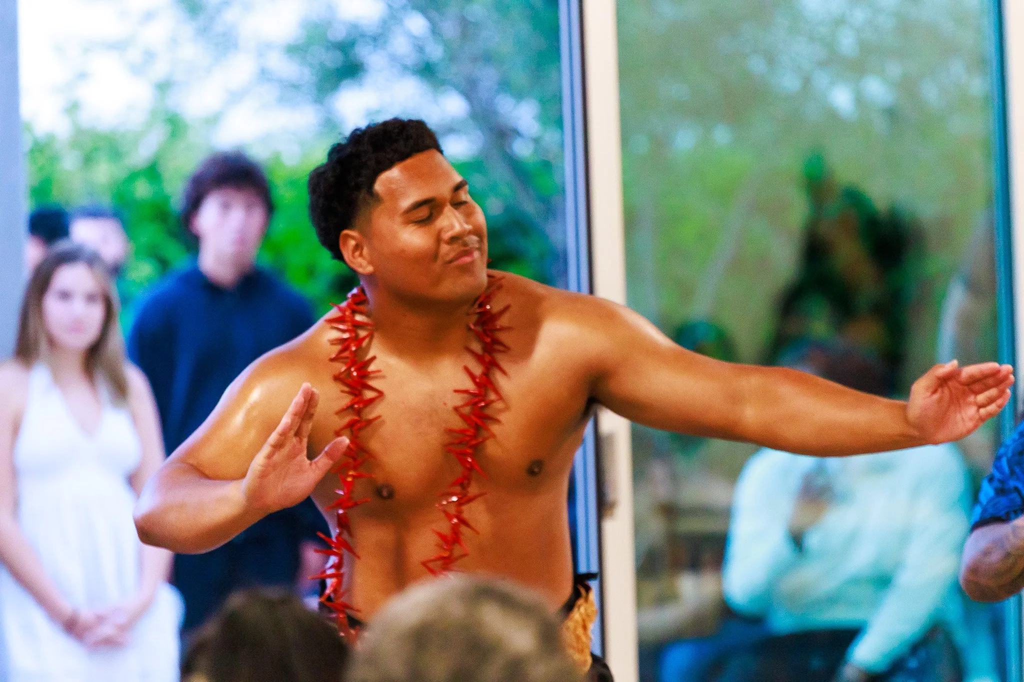 Shirtless man wearing a red chili pepper lei performs a hula-style dance indoors as guests watch.