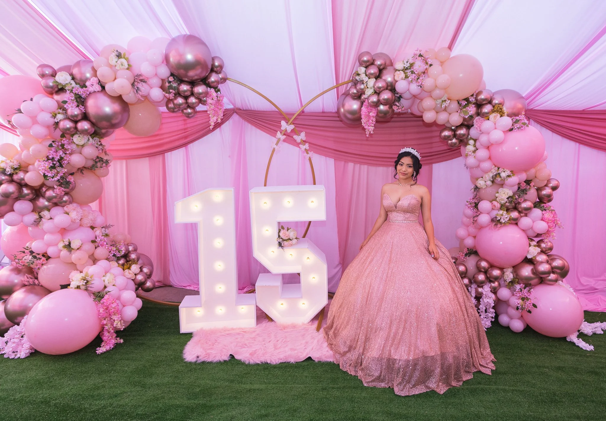 Woman in a pink gown posing beside illuminated number 15 marquee letters amid pink balloon and floral decorations.