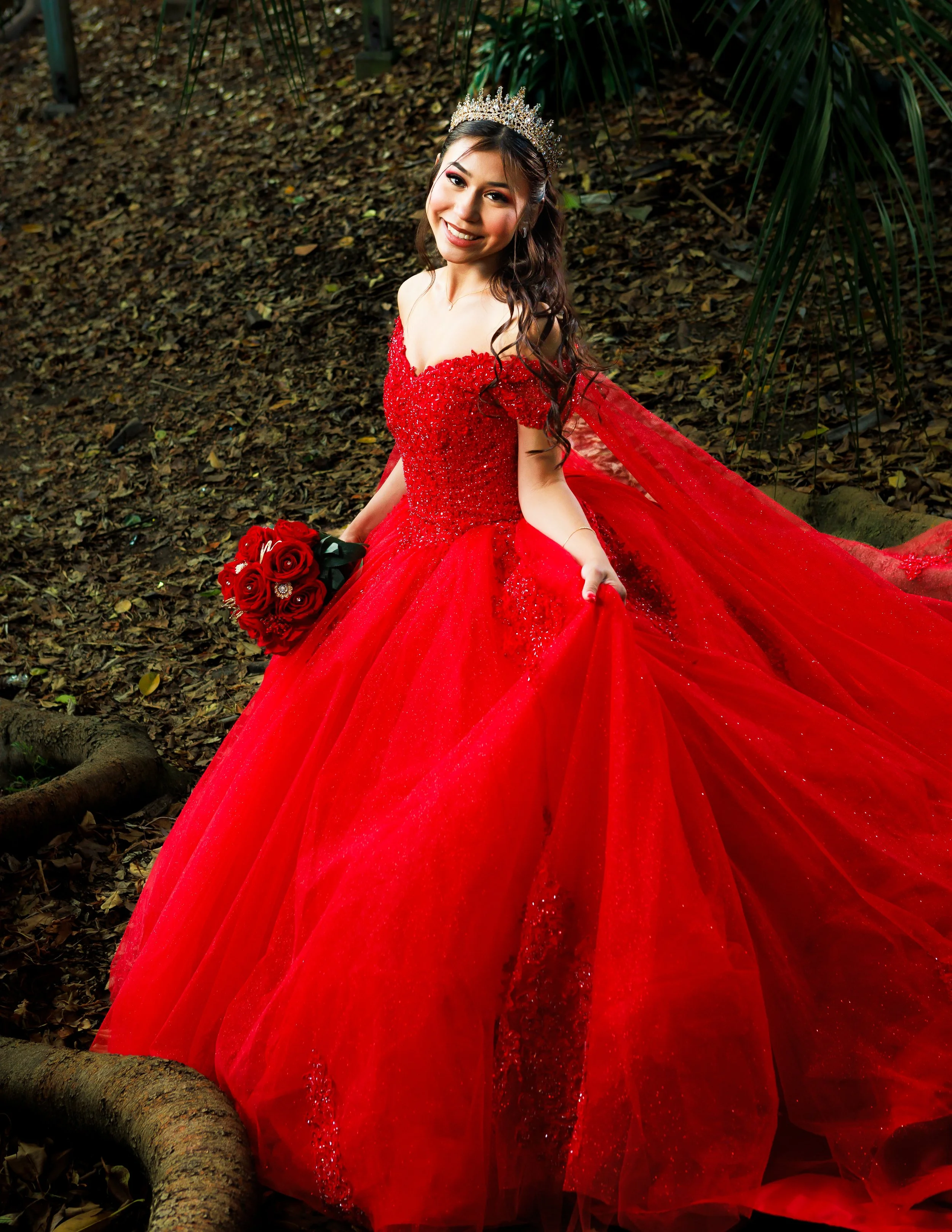 Smiling woman wearing a jeweled tiara and sparkling red ball gown, holding red roses in a forest setting