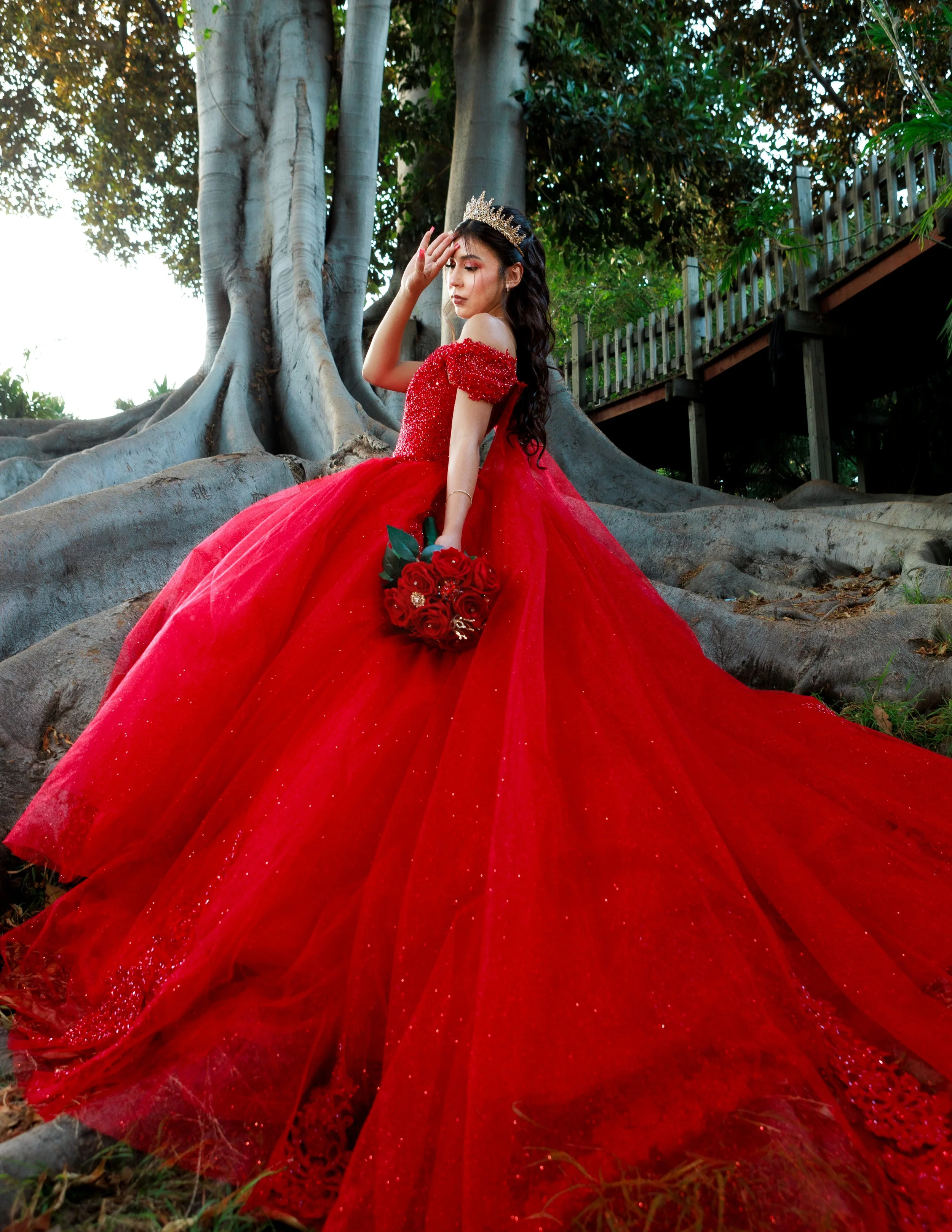 Woman in a sparkling red ball gown and tiara posing beside a large tree with a red rose bouquet.