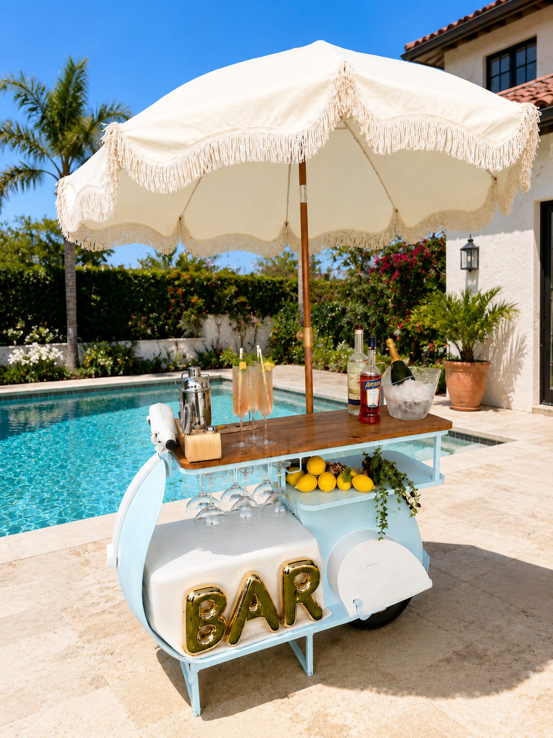 A vintage bar cart with a large white umbrella, set up beside a swimming pool in a sunny backyard. The cart has drinks, lemons, and wine bottles, with a neon "BAR" sign on the front.