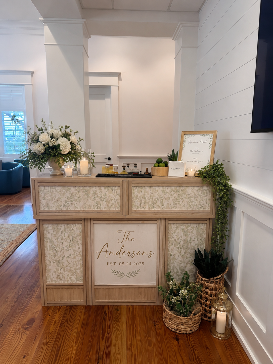Wedding welcome table with floral arrangements, candles, and a sign featuring the family name 'The Andersons' and the wedding date 05.24.2025, in a decorated indoor venue.
