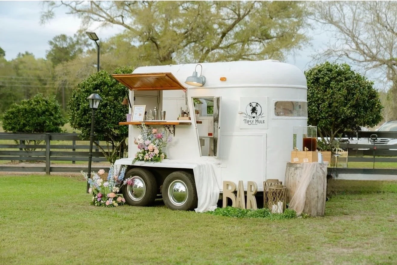 A white vintage trailer set up as a mobile bar with floral decorations and a sign that reads 'Tipsy Mule' on a grassy area with trees and parked cars in the background.