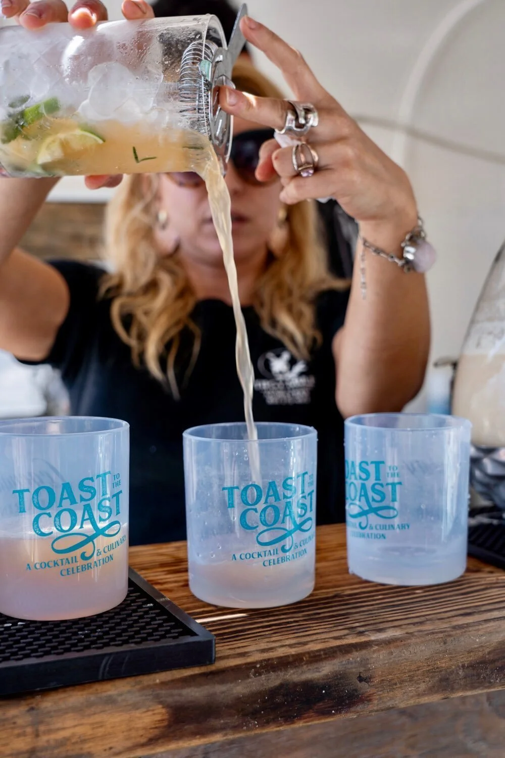 A woman pouring a cocktail from a mixing pitcher into three plastic cups labeled 'Toast to the Coast,' on a wooden bar. She is wearing rings and a bracelet, with a blurred background.