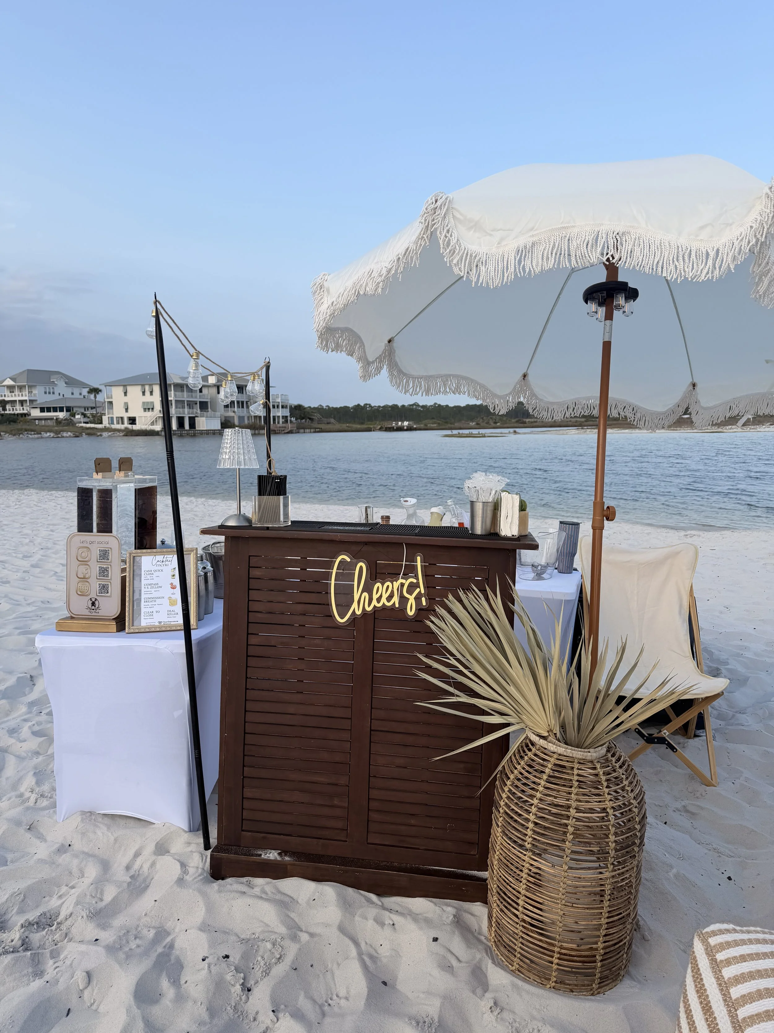 Beachside drink or bar setup with a large white umbrella, wicker basket with dried palm leaves, and a drinks station with a sign that says 'cheers!' on sandy beach, with water and houses in the background.