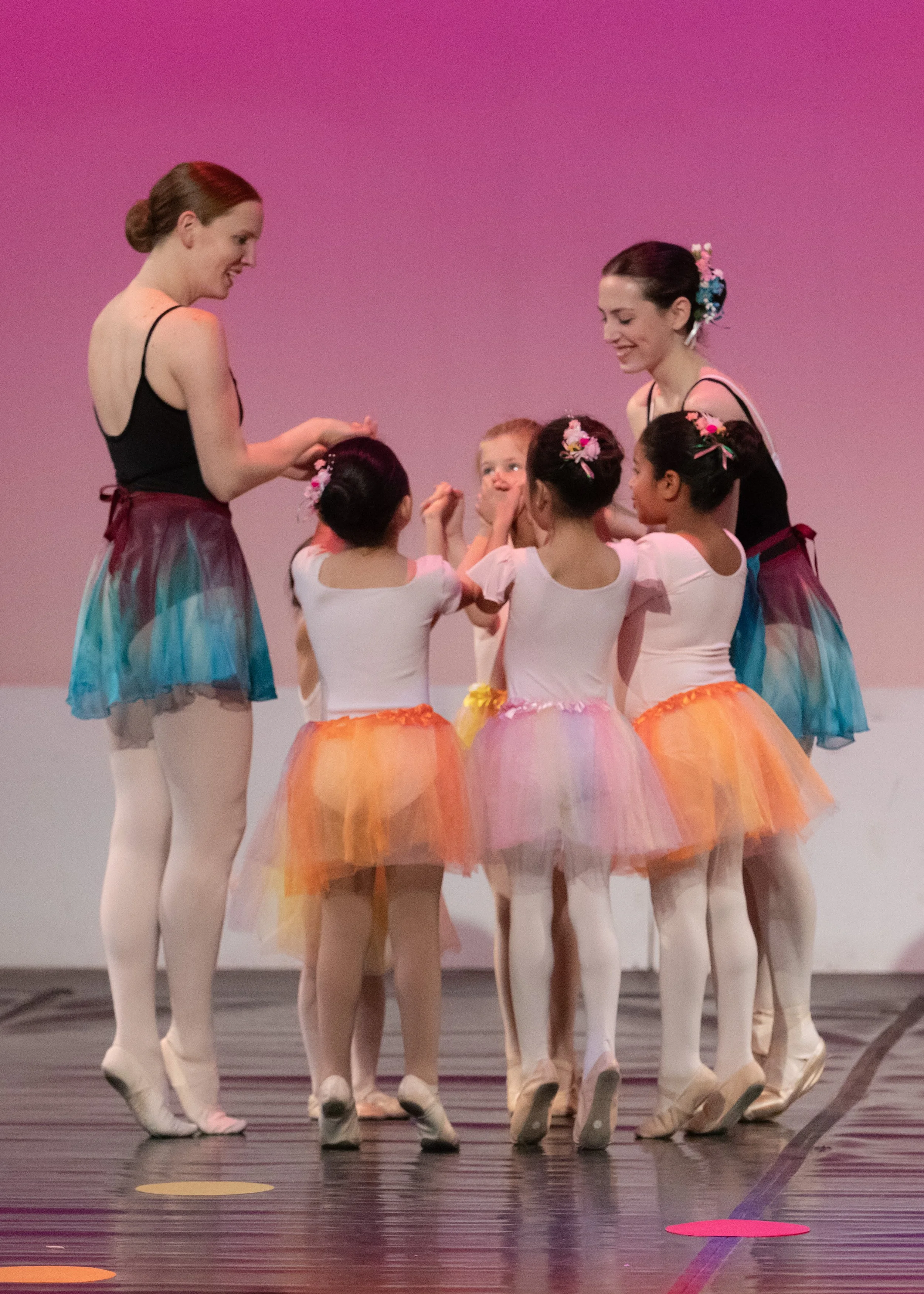 Young dancers and their teachers hold hands in a circle and smile at each other as they stand in a high relevé.