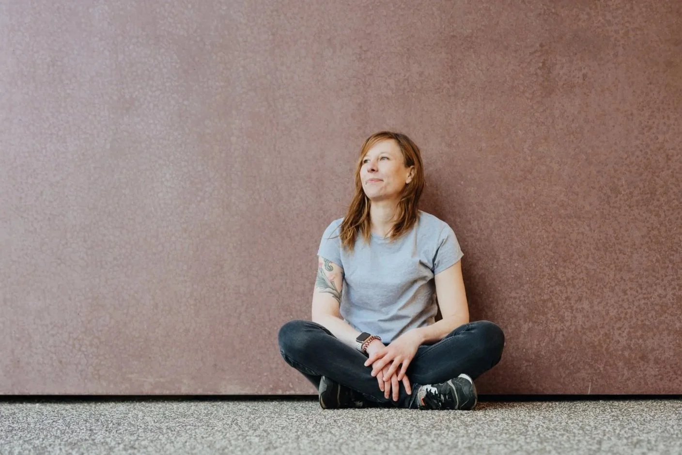 Julia Schmidt sitzt entspannt auf dem Boden vor einer braunen Wand, lächelt leicht und blickt nachdenklich in die Ferne.