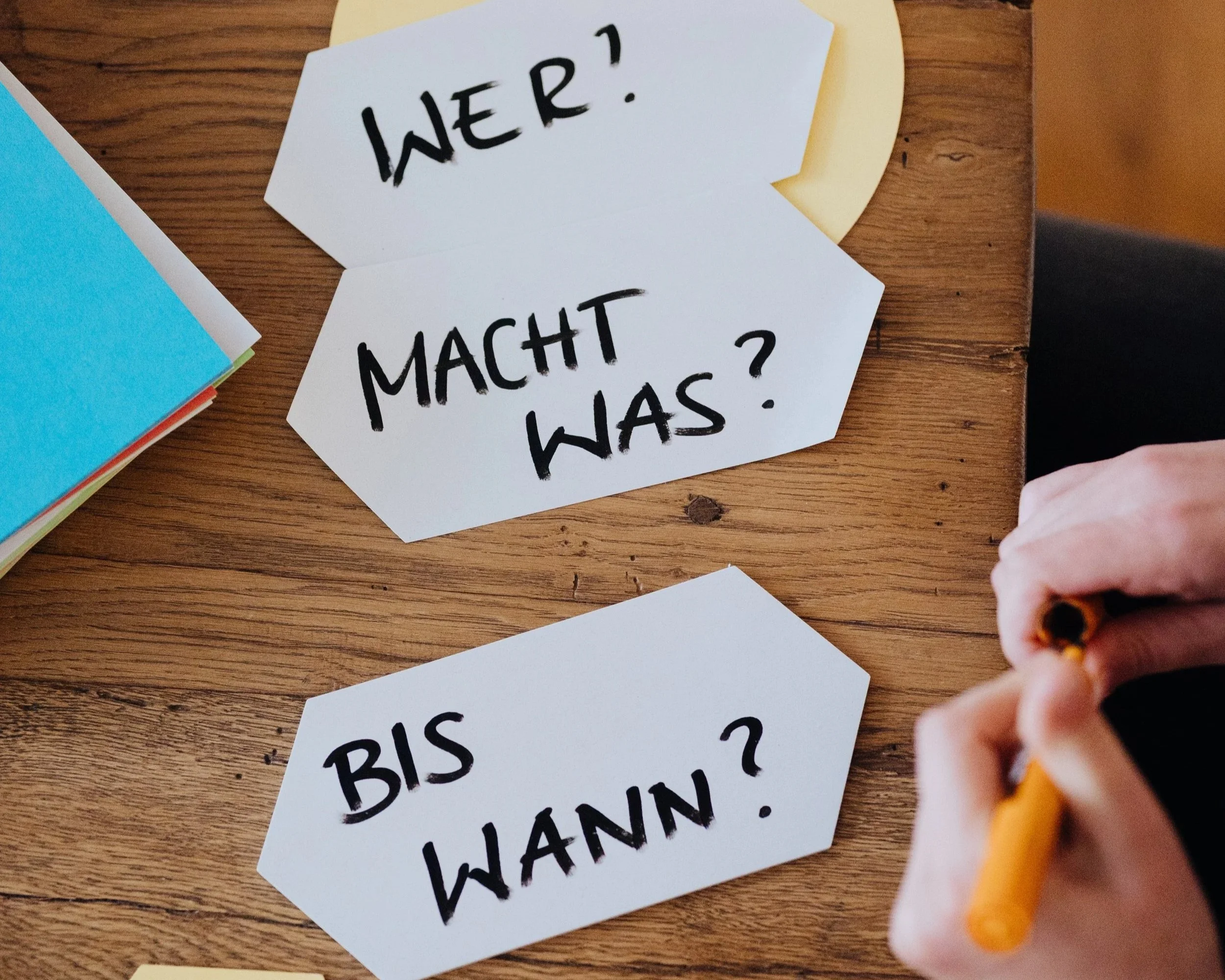 Drei Moderationskarten auf einem Holztisch, die die Fragen 'Wer?', 'Macht was?' und 'Bis wann?' enthalten, während eine Person mit einem Stift schreibt.