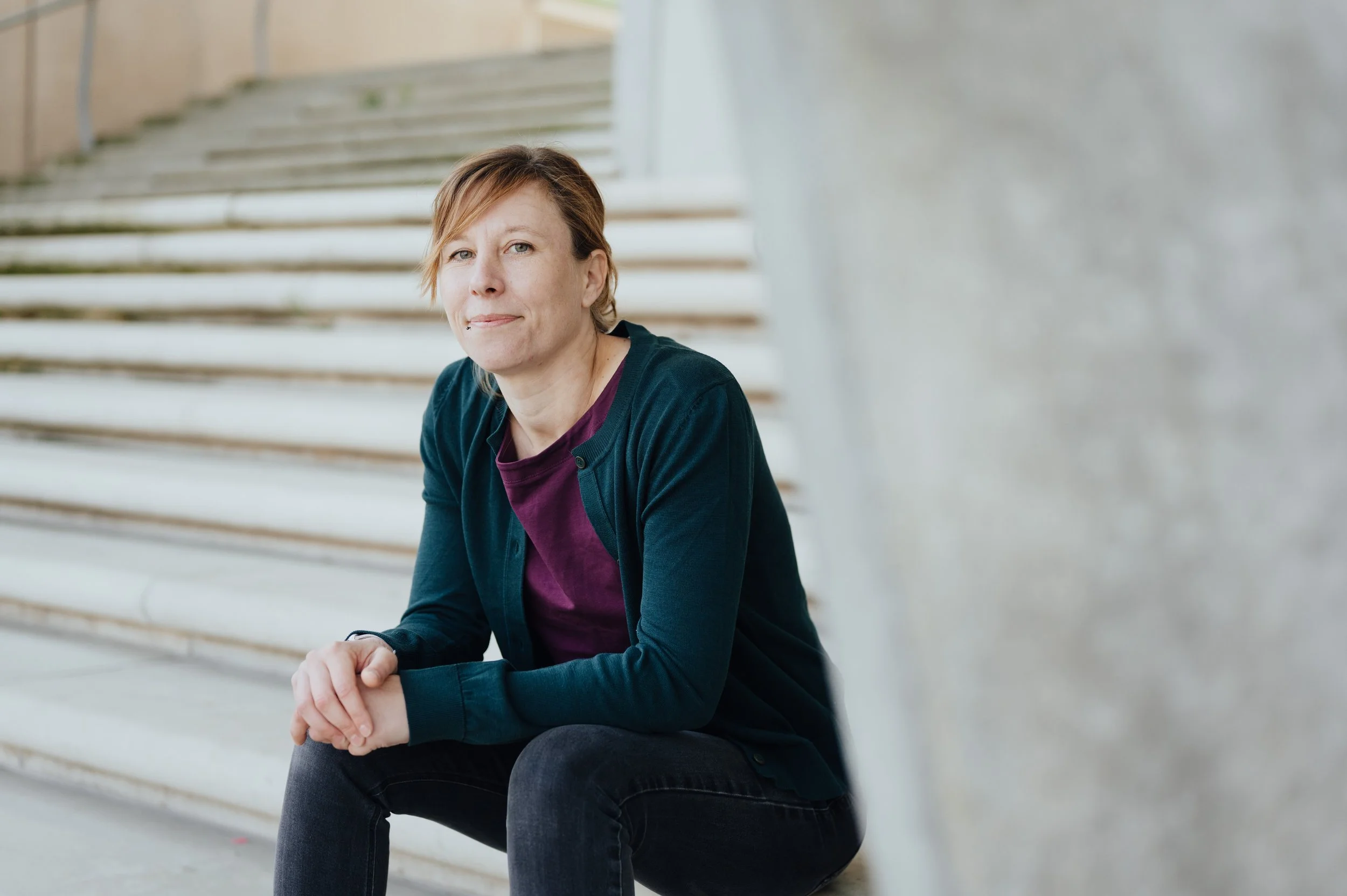 Eine Frau sitzt auf einer Steintreppe. Sie trägt eine petrolfarbene Strickjacke, darunter ein bordeauxfarbenes Shirt. Sie lächelt in die Kamera.