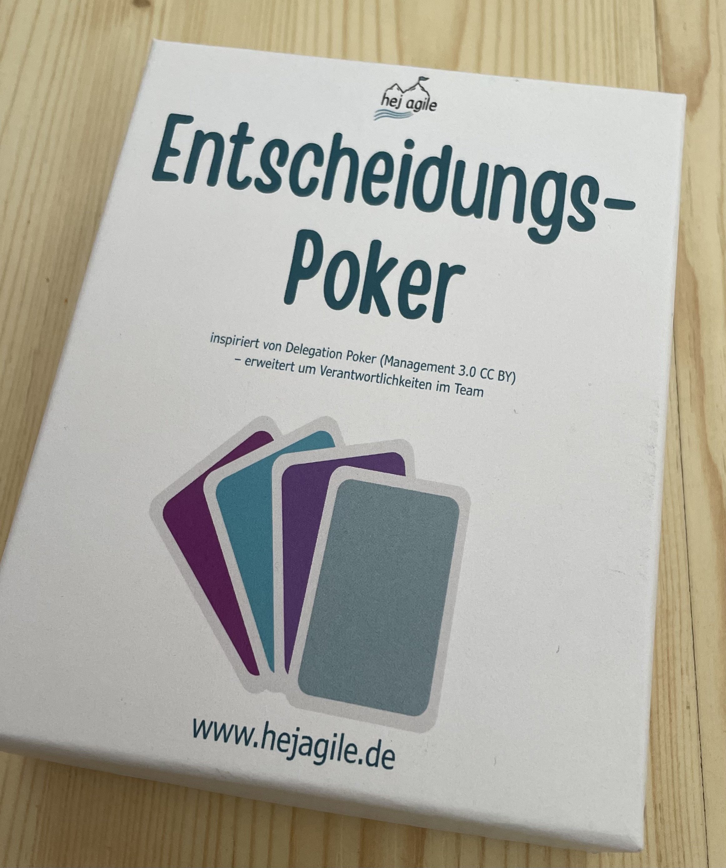 Foto einer weißen Box auf einem Holztisch mit der Aufschrift „Entscheidungs-Poker“ und dem hej-agile-Logo. Darunter sind farbige Karten als Illustration sowie die Webadresse „www.hejagile.de“  zu sehen.