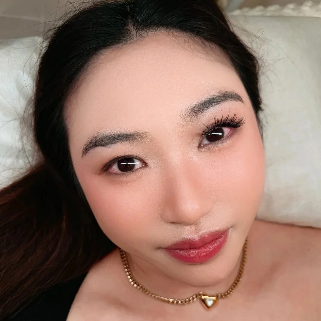 Close-up of a woman with dark hair, makeup, and a gold necklace with a heart pendant, lying on a pillow - fluttery lashes by Pocobeauty.