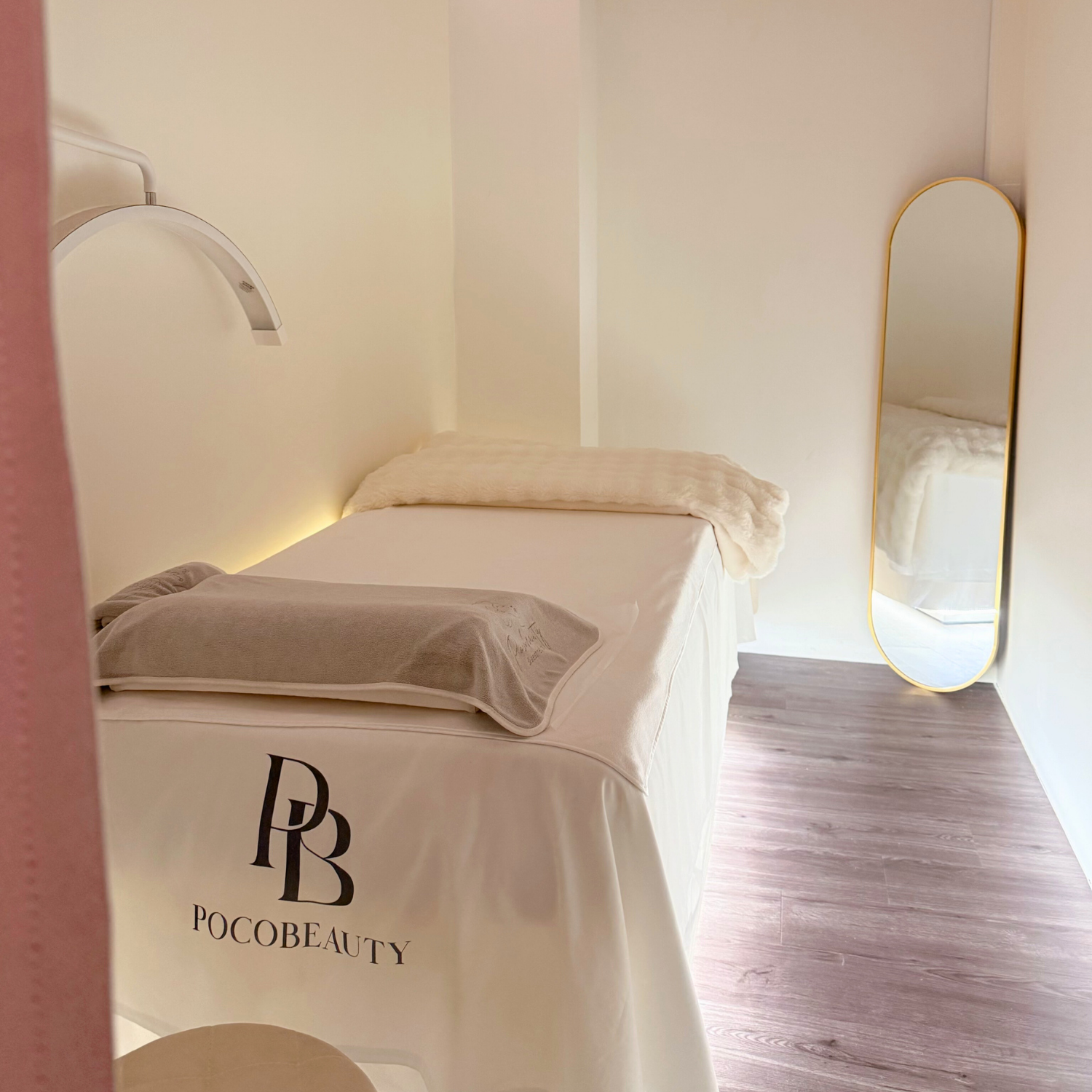 Pocobeauty treatment bed with white linens and a beige towel, positioned in a minimalistic, softly lit room with light-colored walls and a wooden floor. Clean for all your beauty treatments at Chinatown MRT.