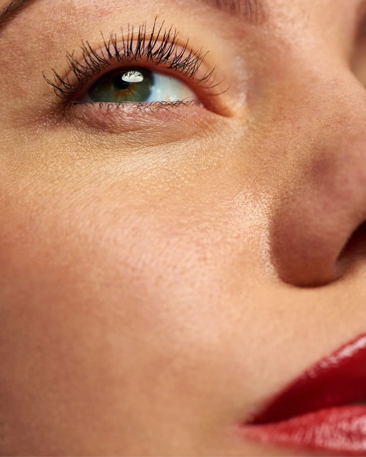 Close-up of a person's eye, showing long eyelashes by Pocobeauty, light brown eye color, and light skin with smooth complexion, partially showing lips with red lipstick.
