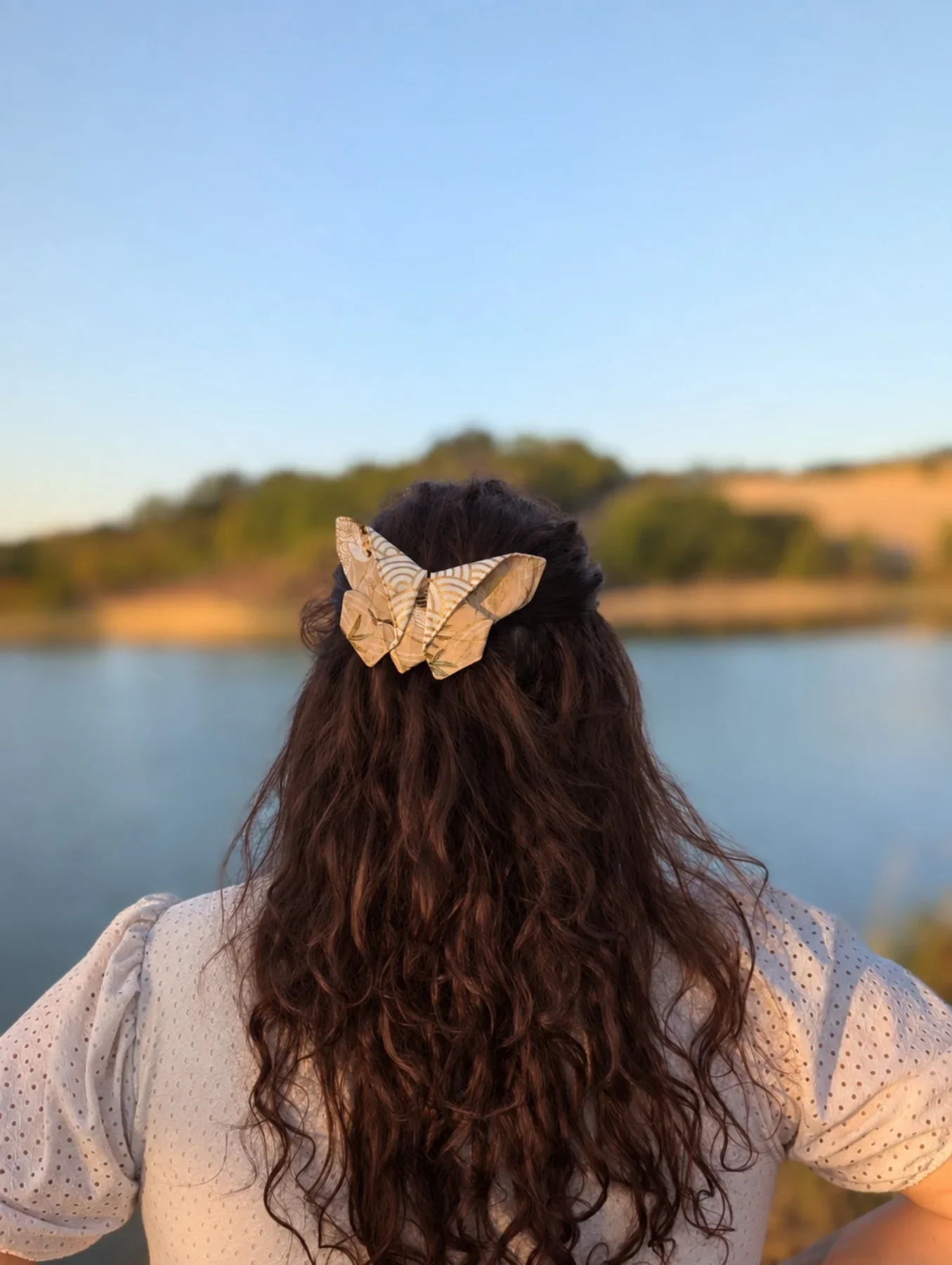 Petit papillon origami ivoire et or porté sur cheveux longs près du lac.