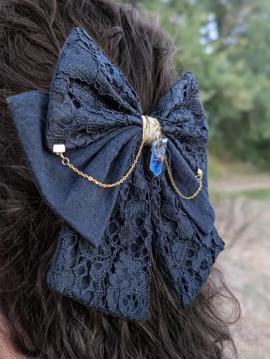 Nœud bijou noir en dentelle, chaînes dorées et cristal irisé sur cheveux bruns, gros plan.