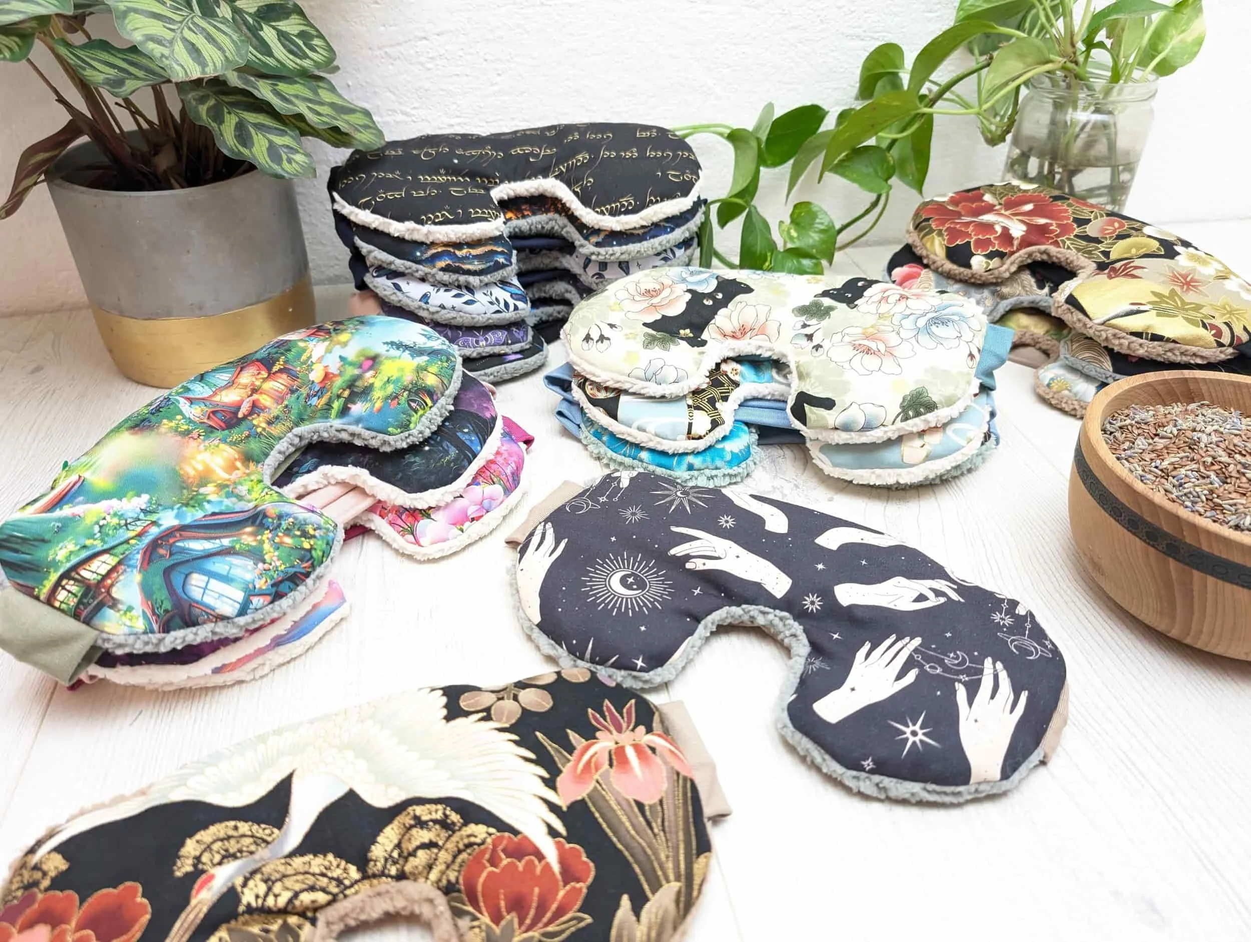 Une collection de masques en tissu colorés et à motifs, disposés sur une table en bois, accompagnés de plantes et d'un bol contenant des graines.