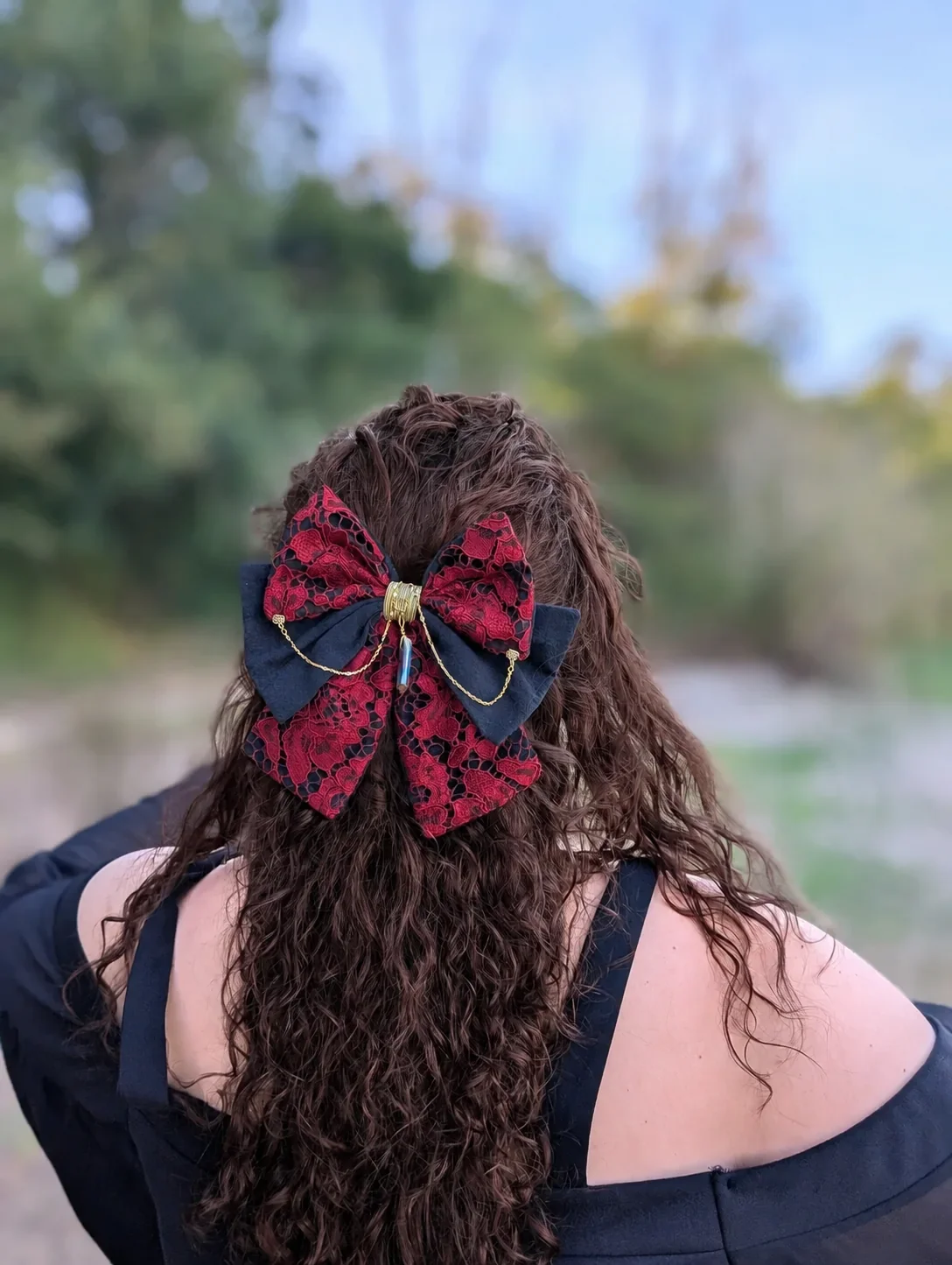 Noeud bijou rouge et noir en dentelle avec chaînes dorées, porté sur cheveux bouclés.