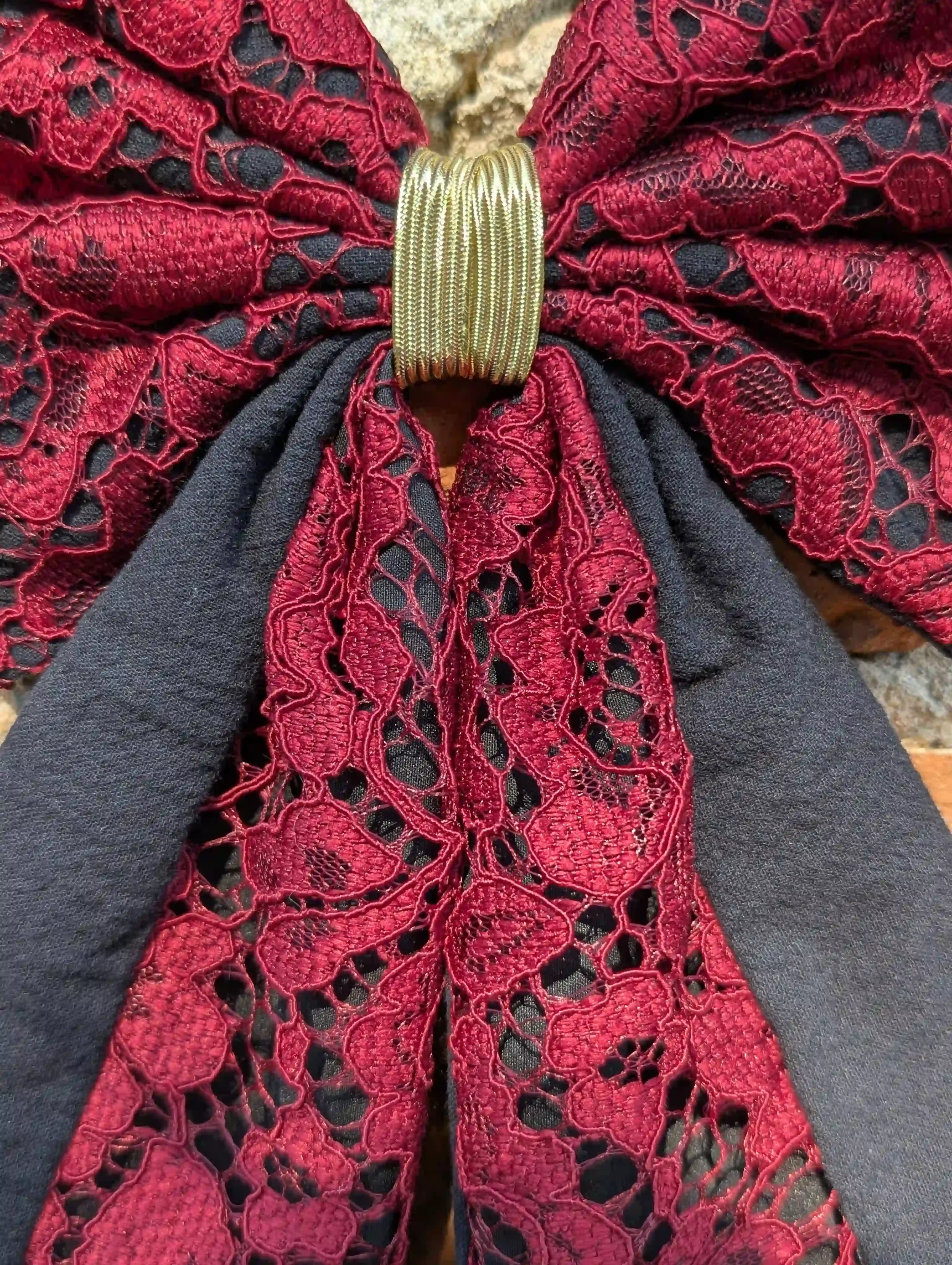 Détail d’un noeud pour cheveux en dentelle rouge profond et tissu noir avec finition dorée au centre.