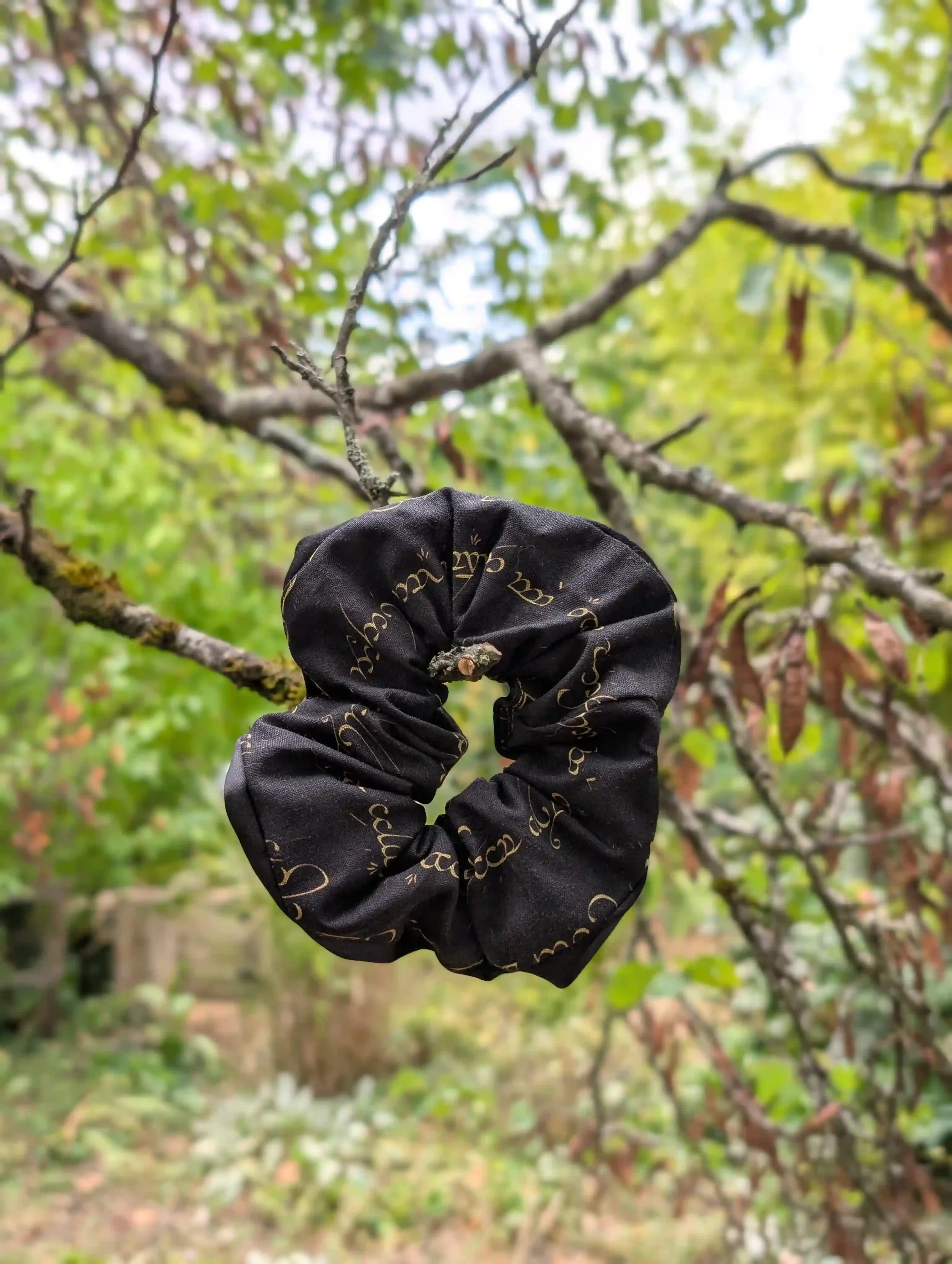 Grand chouchou Elfique noir et or avec satin noir, présenté suspendu dans un décor végétal