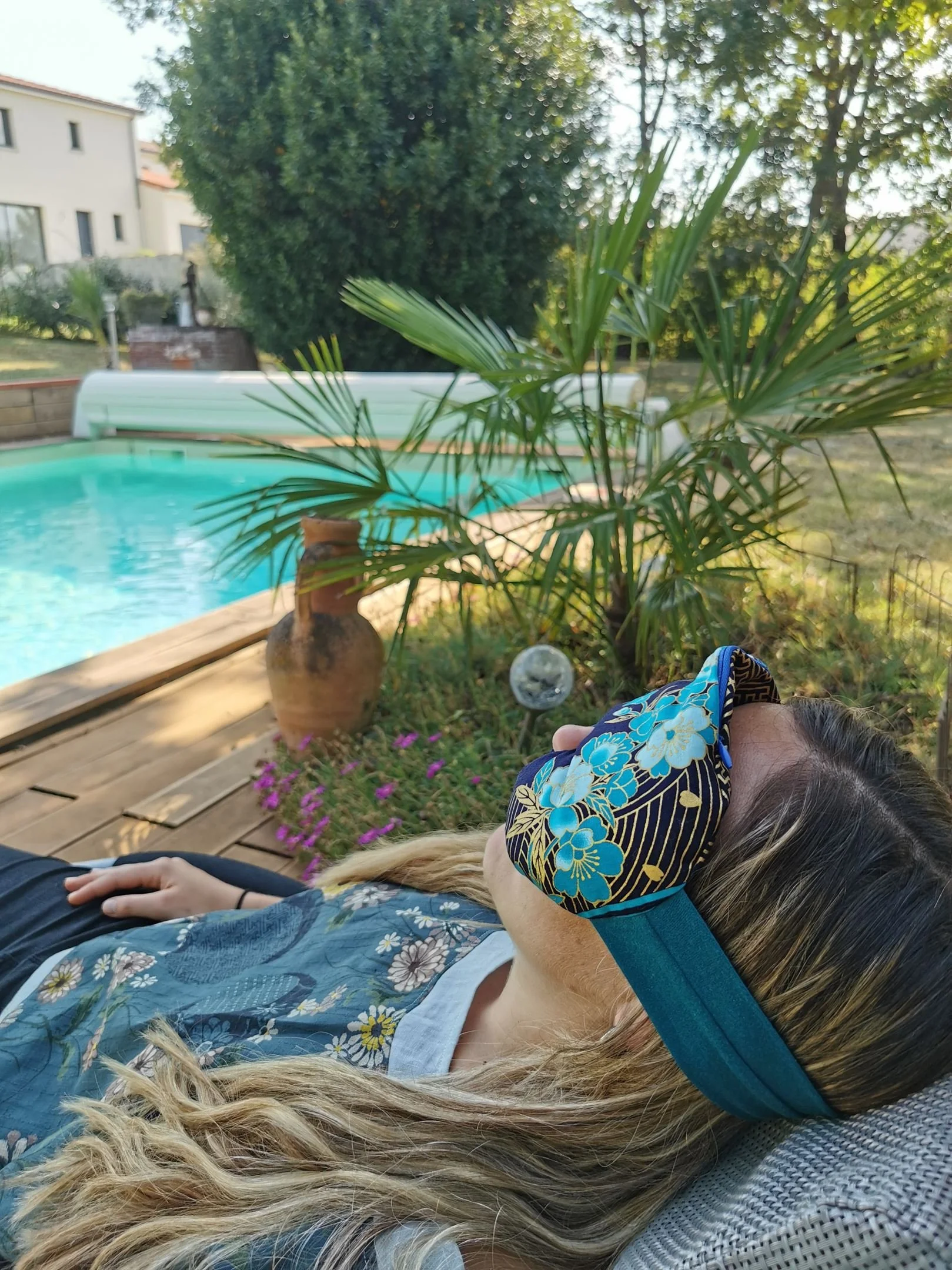 Une femme avec un masque de sommeil floral repose dans un jardin ensoleillé, près d'une piscine, avec des plantes et des bâtiments en arrière-plan.