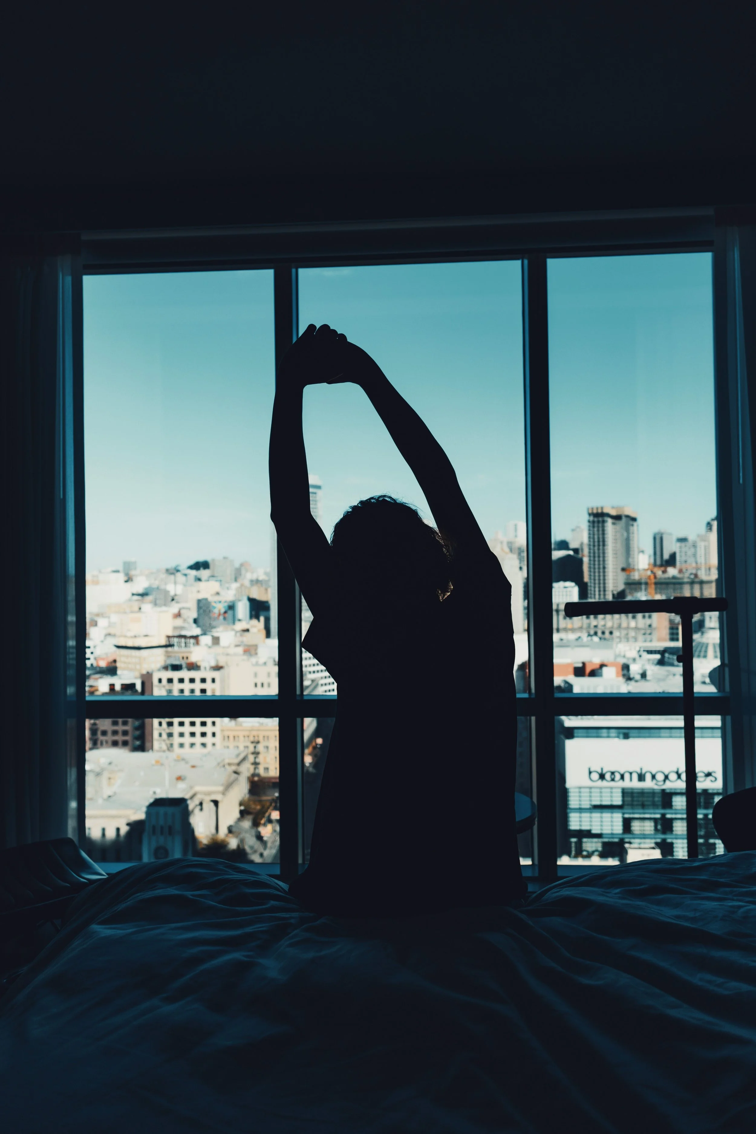 Silhouette d'une femme s'étirant devant une grande fenêtre au réveil, dans une chambre baignée de lumière naturelle.