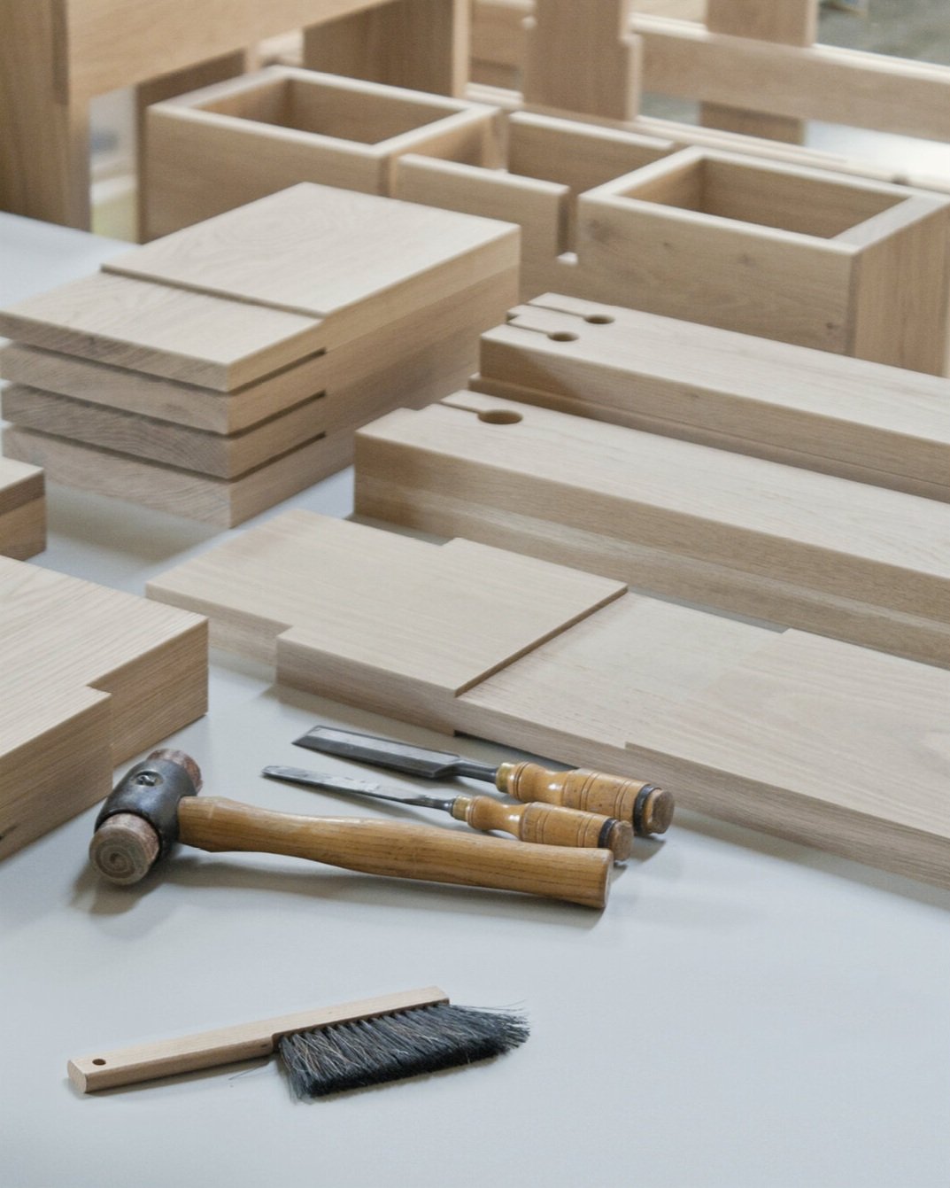 Various unfinished wooden components of By Dave's tables, tools including a hammer and chisels, and a small brush on a workspace, likely for woodworking or carpentry.