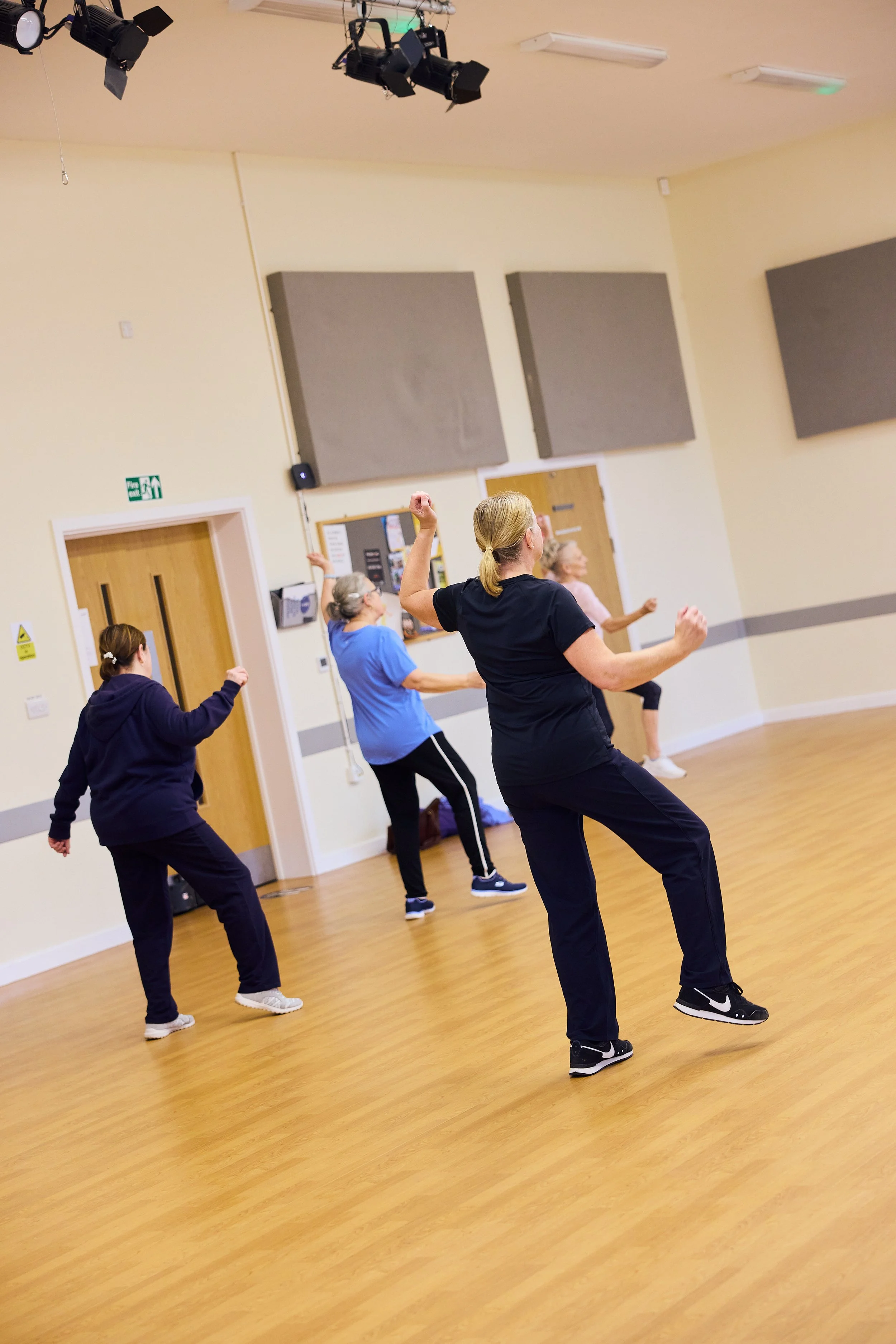 A group of women participating in a dance or exercise class in a studio with wooden floors and beige walls. They are moving with their arms raised or extended.