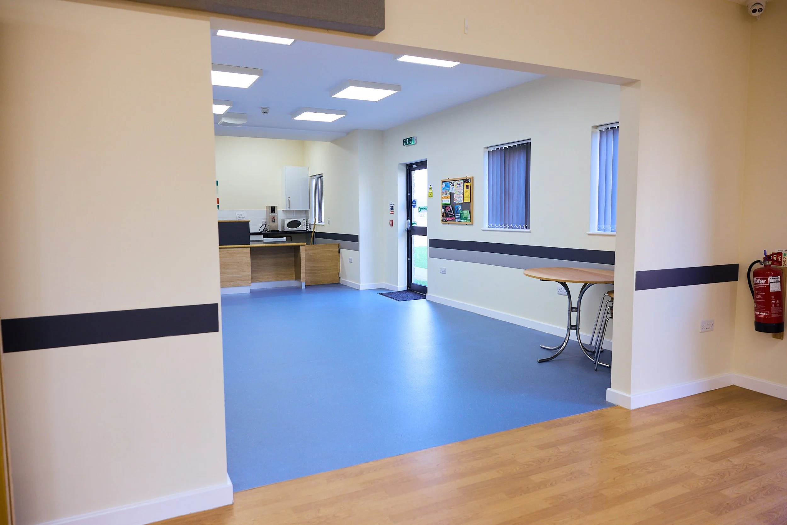 View of an empty room with light yellow walls, blue flooring, and white and wood accents, including a kitchenette area with a microwave and cabinets, two large windows with vertical blinds, a bulletin board, a small round table with stacked chairs, and a red fire extinguisher mounted on the wall.