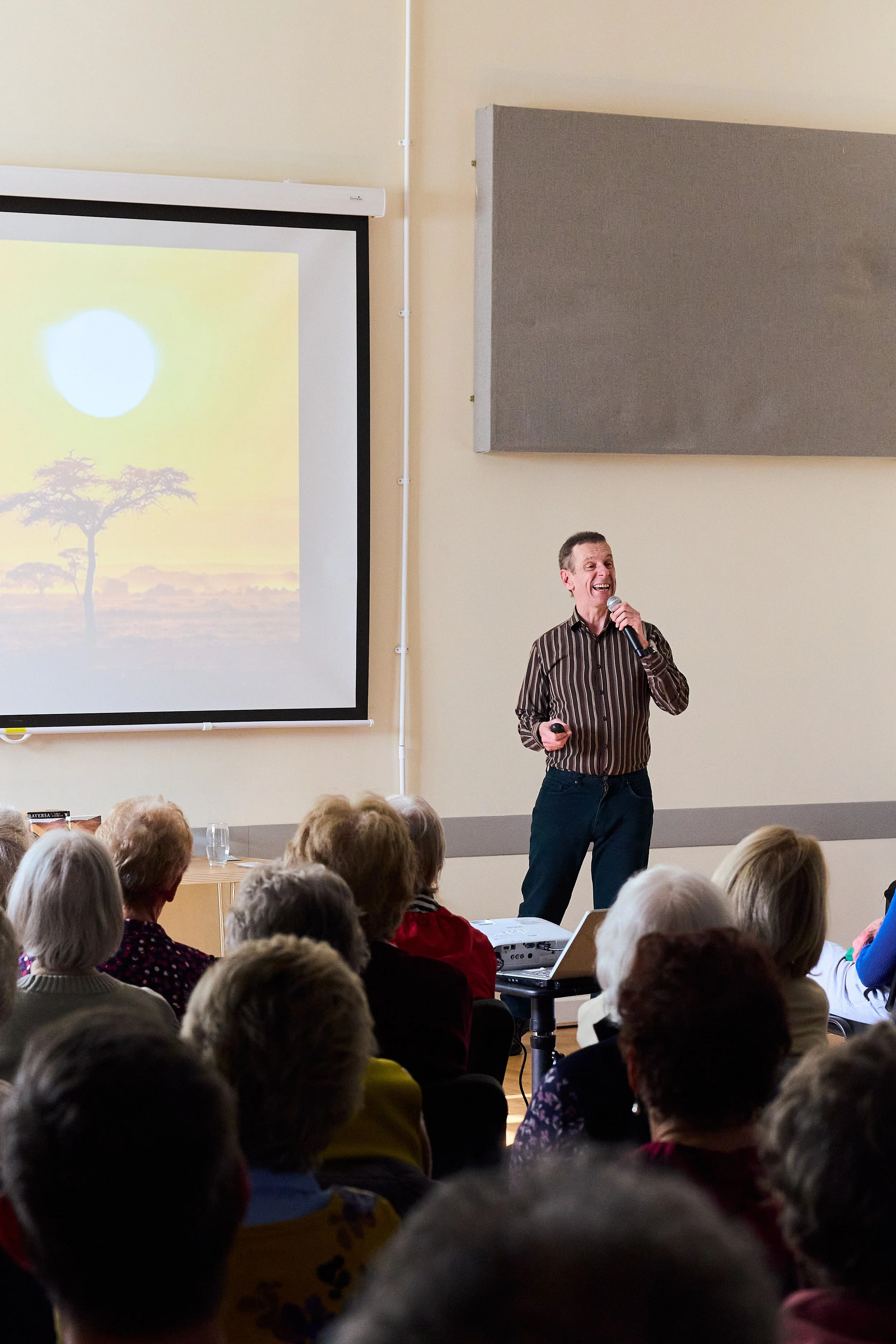 A man giving a presentation to an audience in a room, with a slide showing a sunset and trees projected on a screen.