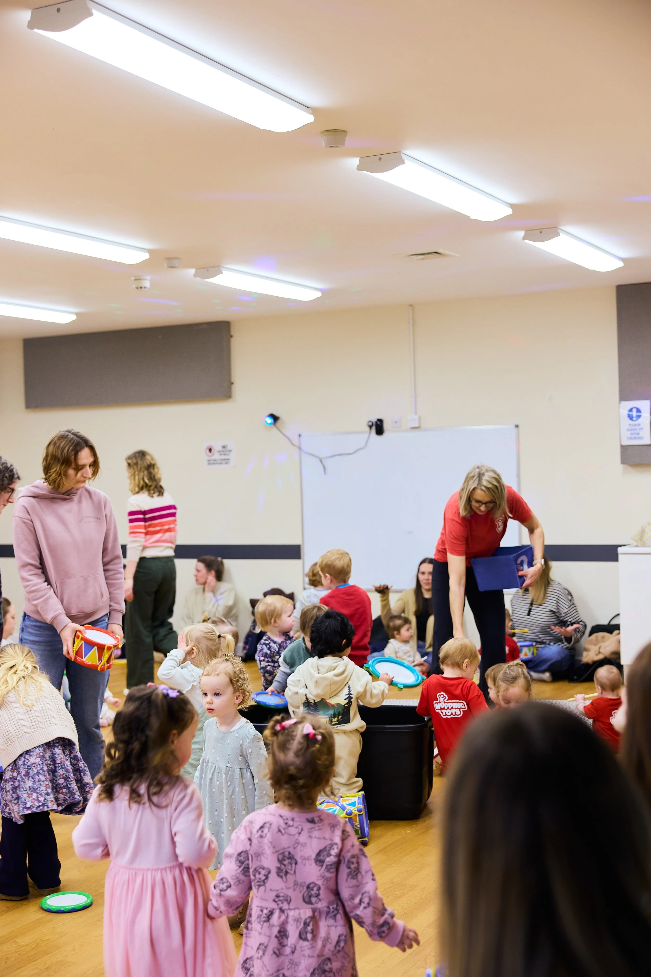 Children gathered in a room with some adults, sitting and standing, participating in an activity, with a whiteboard and a small sign on the wall.
