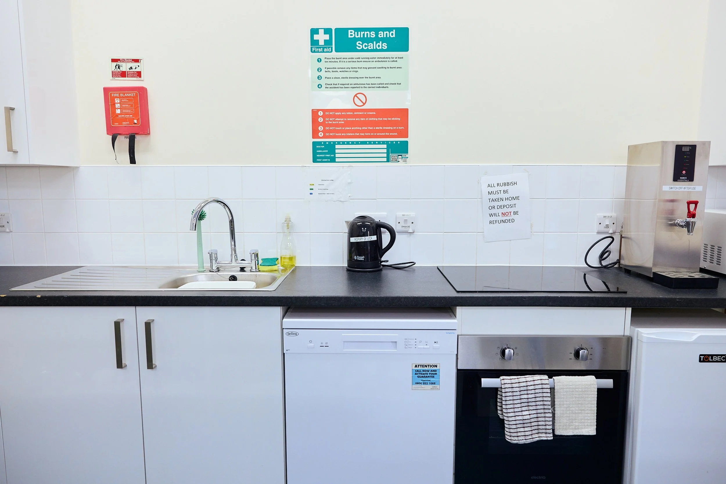 A kitchen area with a white sink, black countertop, and white cabinets. There is a dish soap bottle, a black kettle, a dryer, and a small white fridge. Signs on the wall include burn and scalds warnings and rules about rubbish disposal. A fire blanket is mounted on the wall.