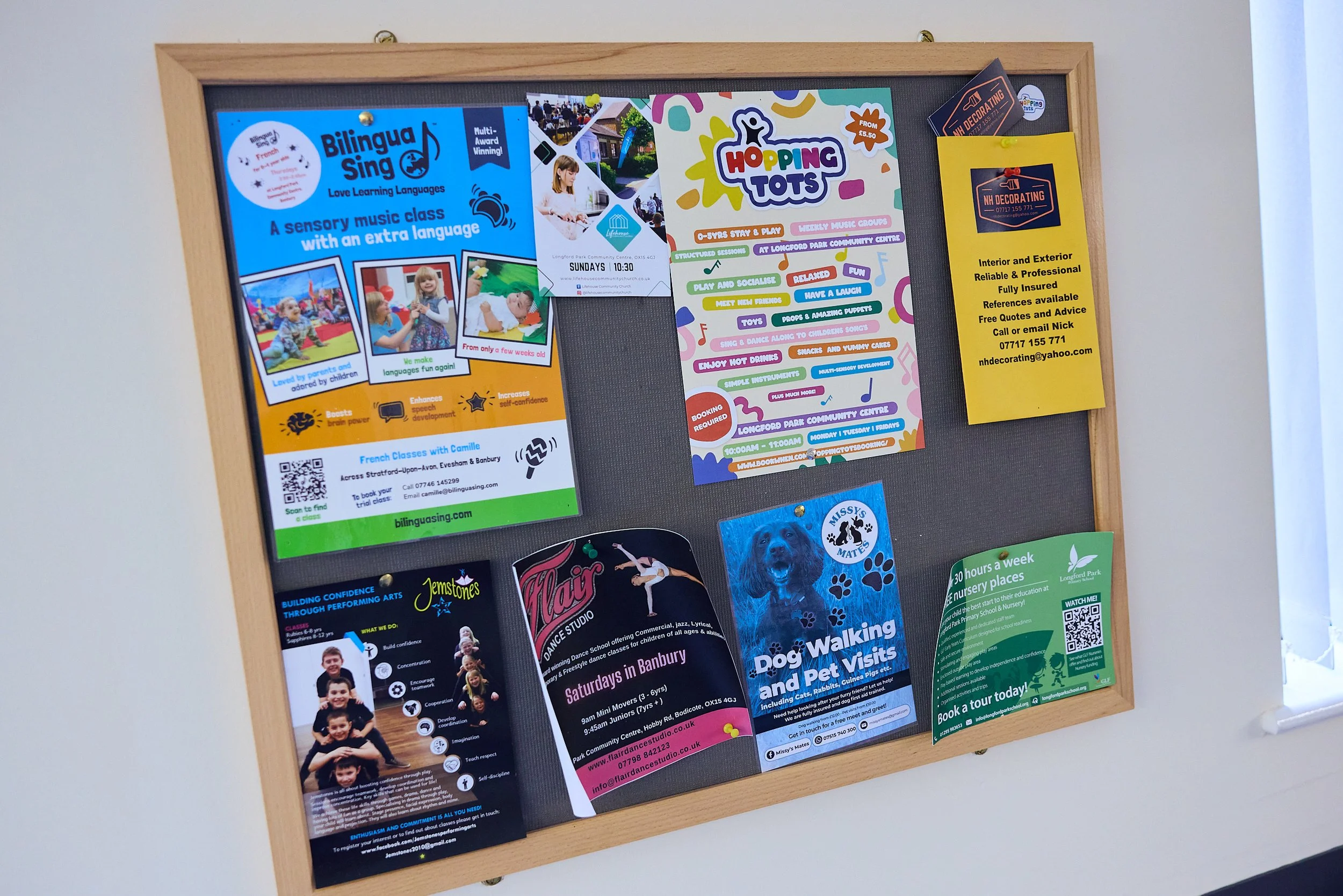 A bulletin board with various colorful flyers and posters advertising classes, activities, and services including bilingual singing classes, hopping tots, dog walking and pet visits, and nursery places. The board has a wooden frame and is mounted on a white wall.