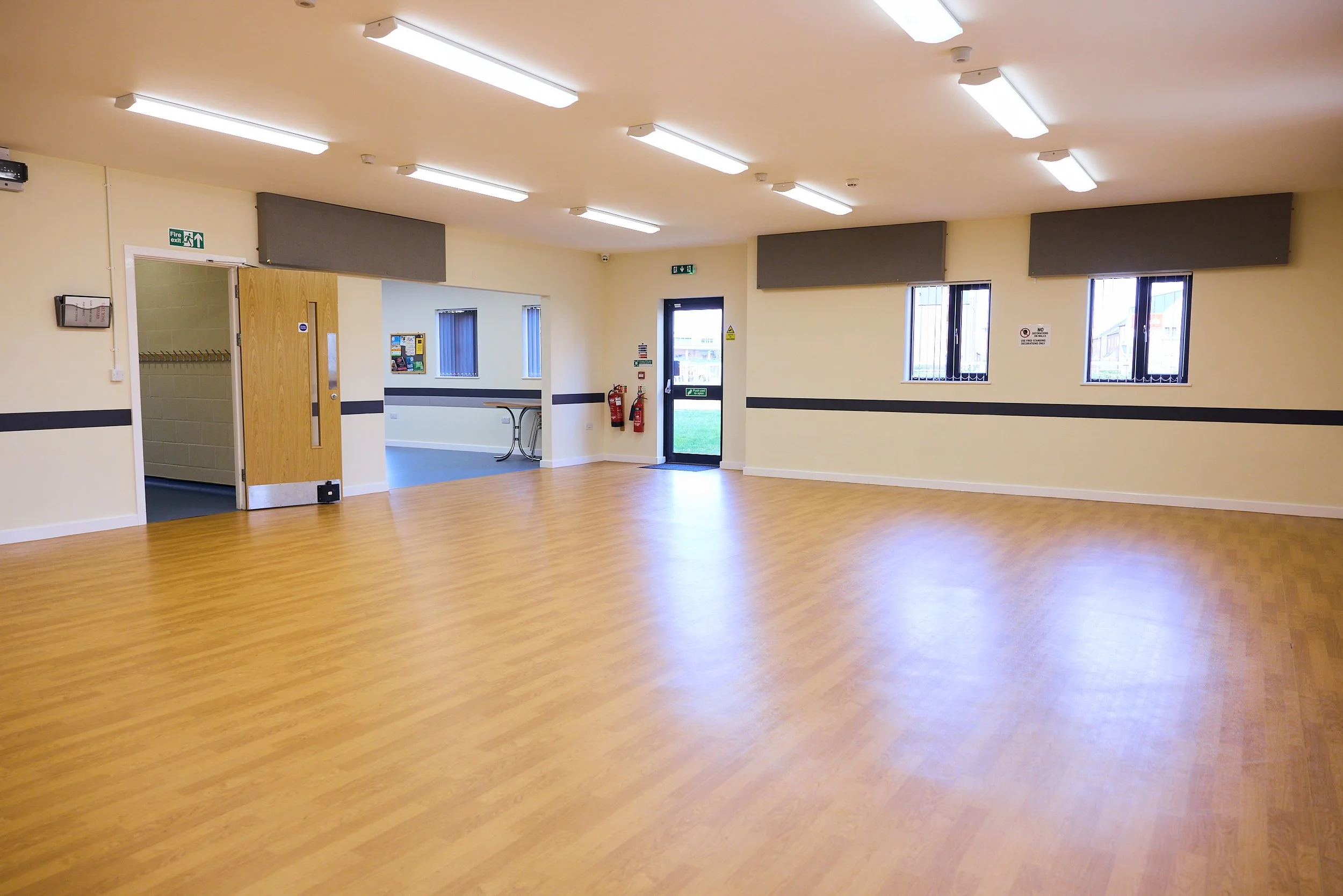 Empty room with wooden laminate floor, beige walls, fluorescent ceiling lights, and windows with bars. There is a door leading outside and fire extinguishers on the wall.
