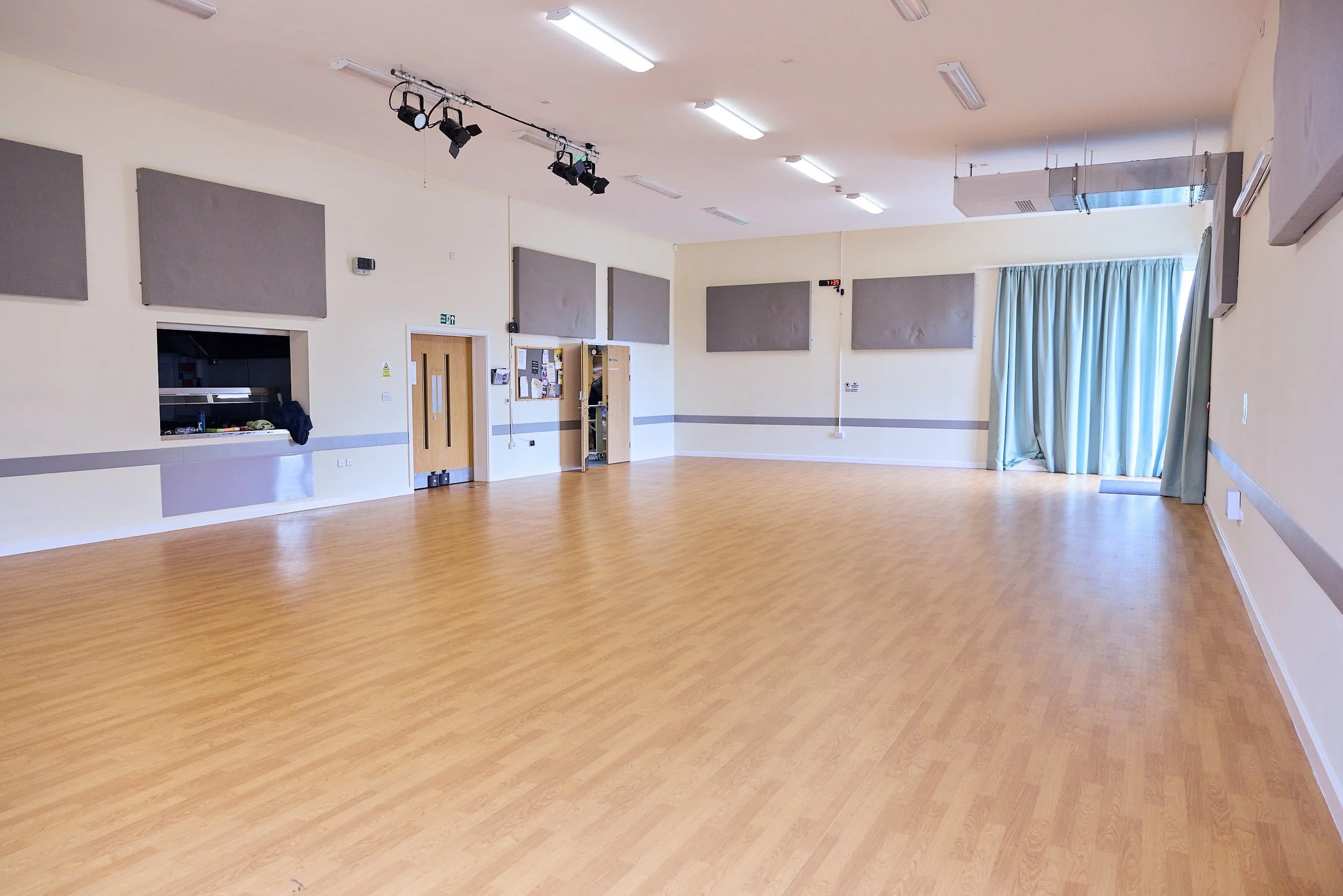 Empty multi-purpose room with wooden flooring, beige walls, gray acoustic panels, a window with a blue curtain, and a door with bulletin boards.