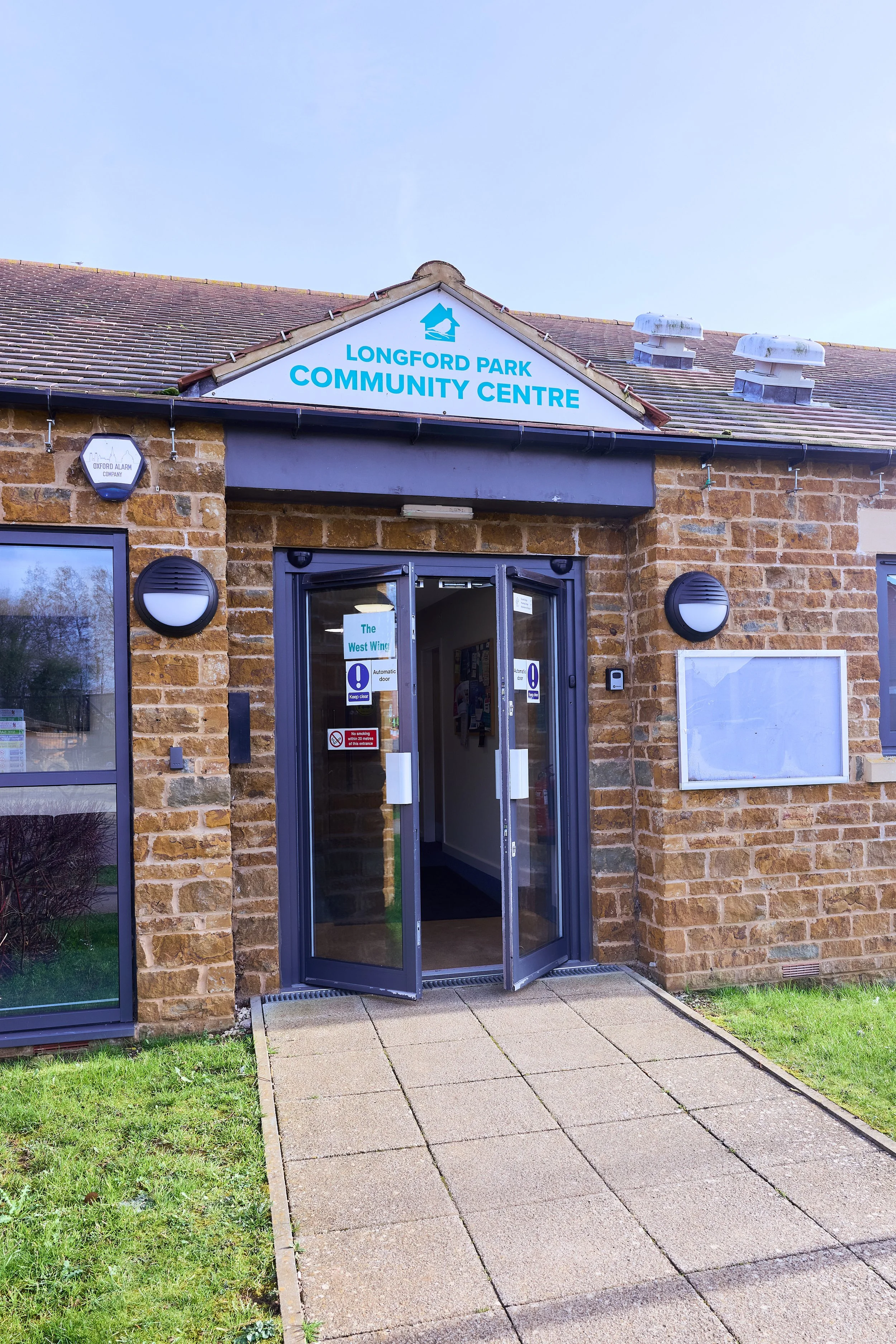 Brick community center building with a sign reading 'Longford Park Community Centre,' glass double doors at the entrance, and a concrete walkway leading inside.