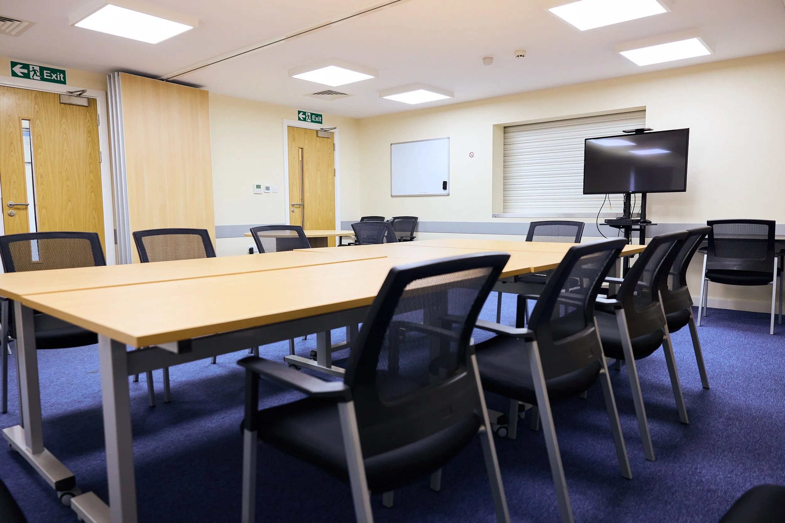 Empty conference room with a U-shaped table, black chairs, a whiteboard, a television, and exit signs.