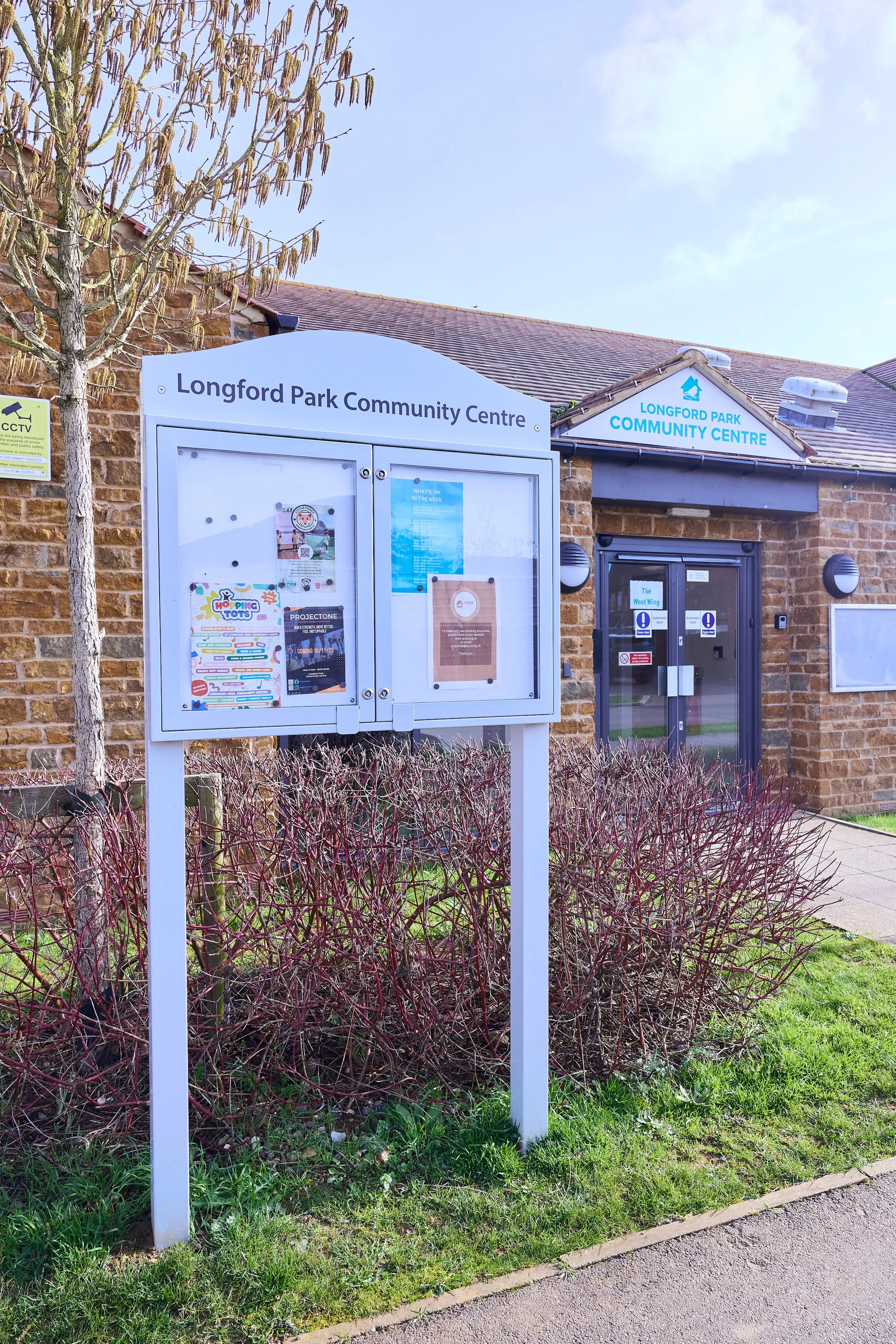Signboard at Longford Park Community Centre, with posters and notices, in front of the brick building with a sign reading 'Longford Park Community Centre' and a glass door.