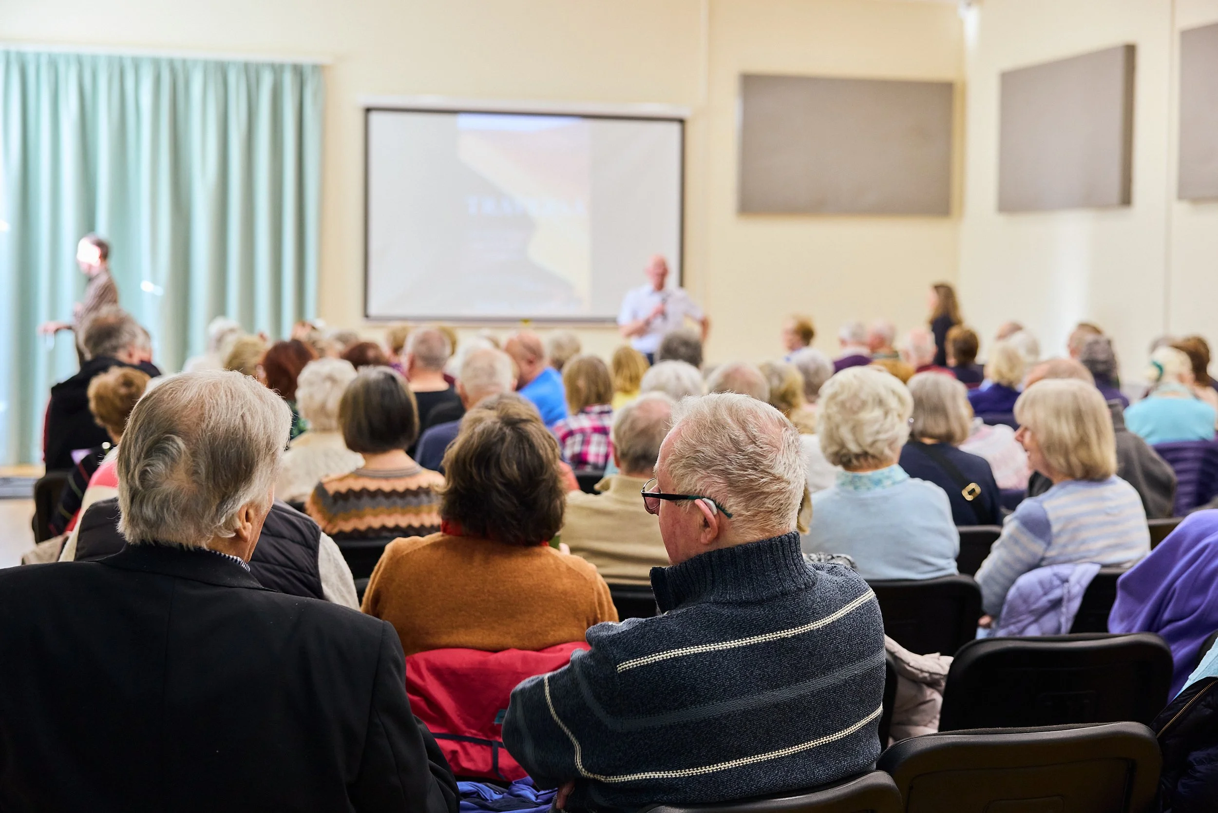 A large gathering of elderly people seated in a room attending a presentation or lecture. A speaker stands in front of a projection screen, and a woman stands nearby. The room has light-colored walls and curtains.