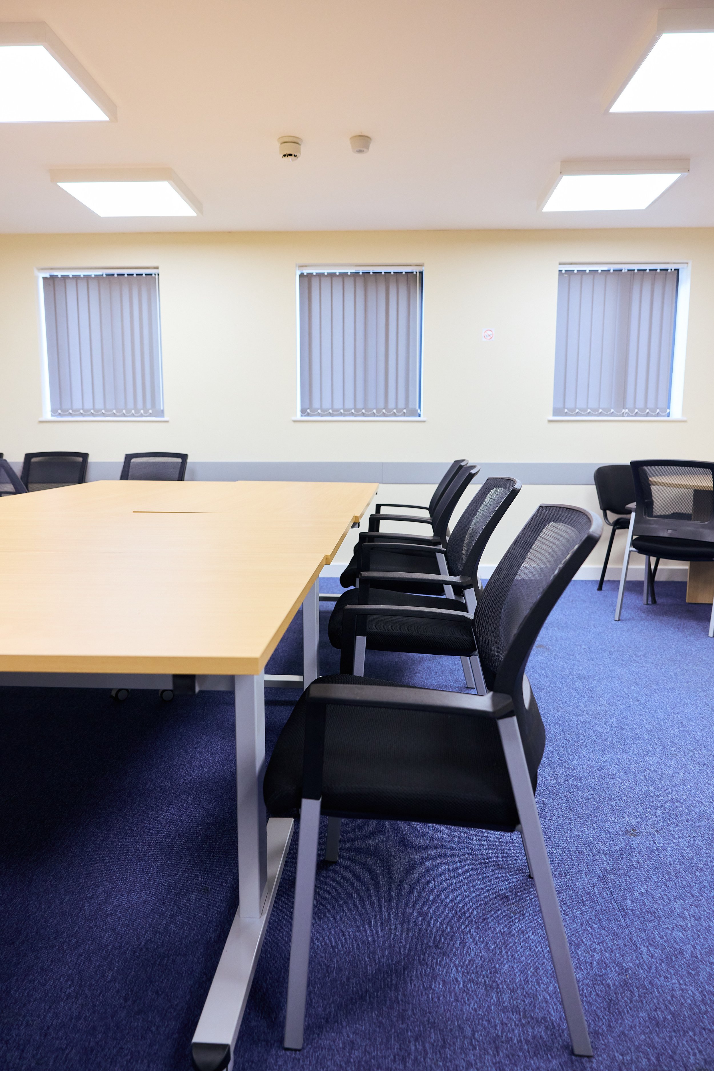 Empty conference room with rectangular table and black chairs, three windows with closed blinds, blue carpet, and ceiling lights.
