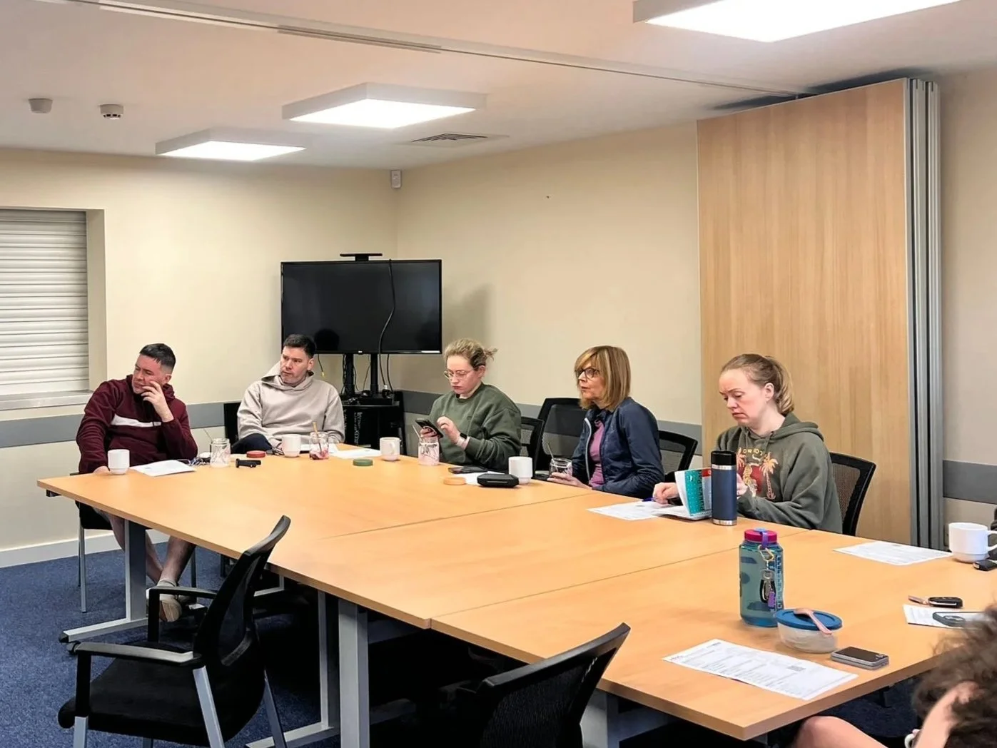 Person gesturing with hands in front of a laptop with a presentation slide on the screen, a notebook, and a phone on a wooden conference table, with other people working nearby.