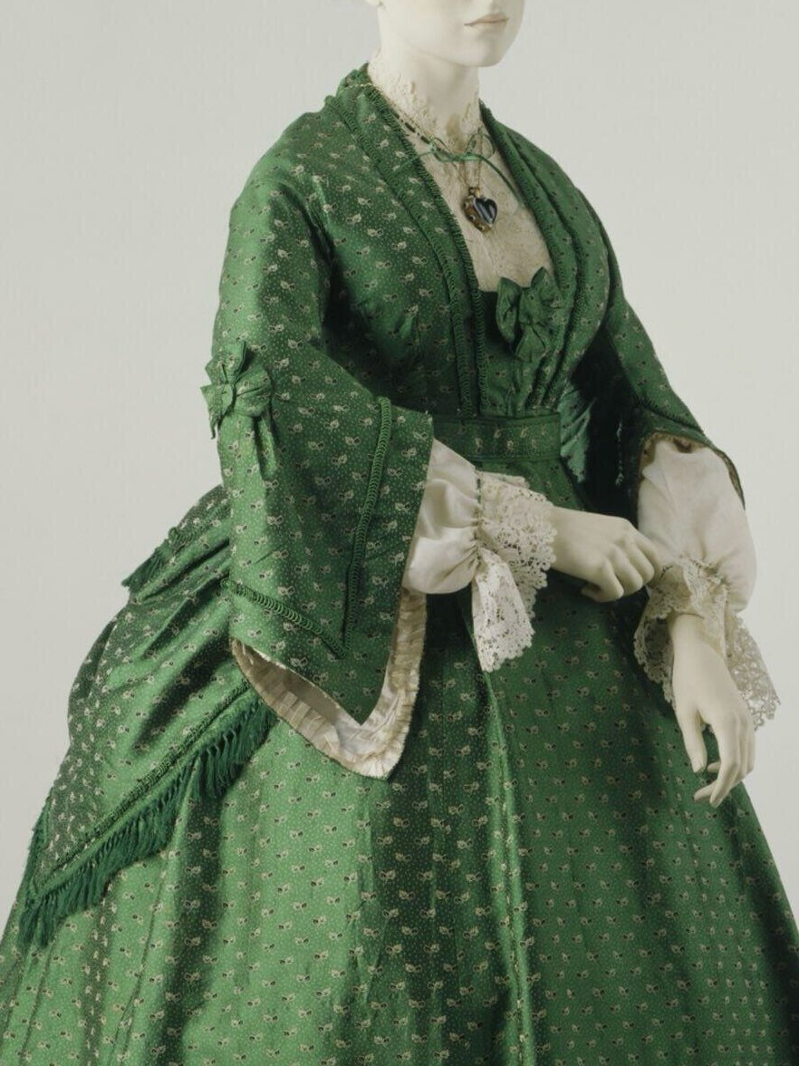 💚 &ldquo;This ensemble is typical of fashionable women&rsquo;s daywear of the early 1870s. The silk has a figured pattern of black and white leaves on a speckled ground. The dress is trimmed with silk braid. There is an overskirt over the back of th