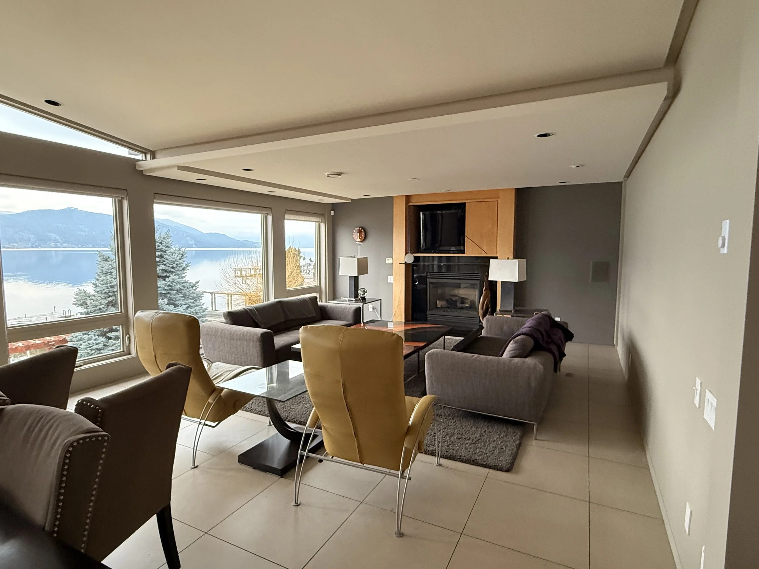 Interior painting project in Vernon, BC. Living room with large windows overlooking a lake and mountains, furnished with sofas, chairs, a glass coffee table, and a fireplace.
