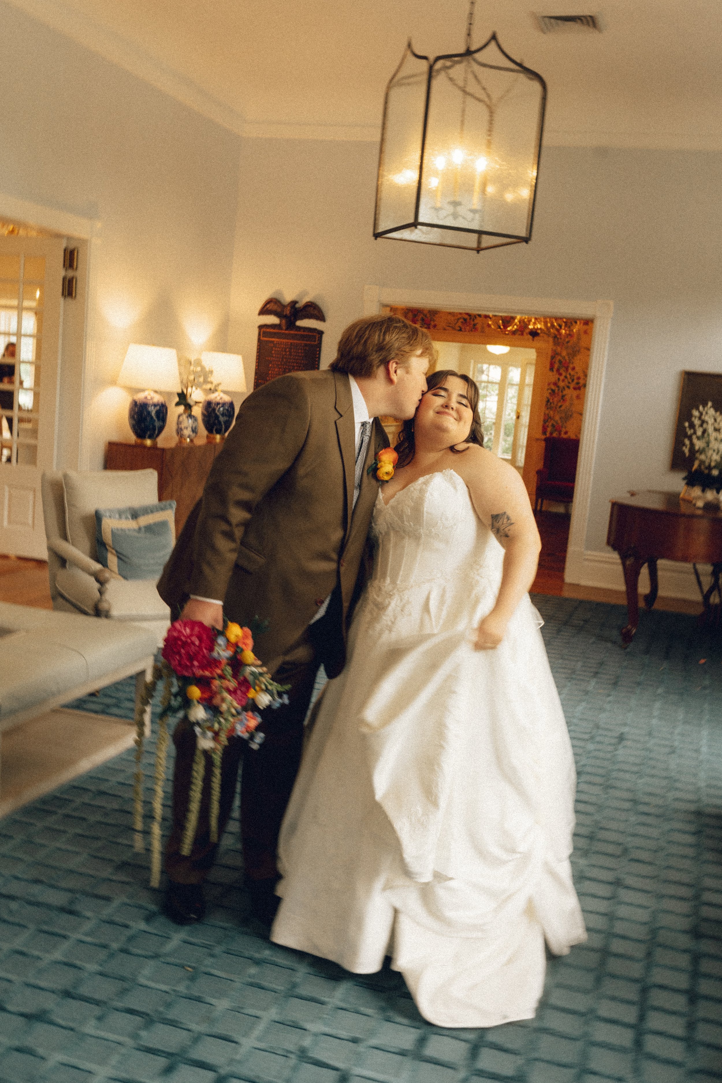 A bride in a white wedding gown smiling with closed eyes, being kissed on the forehead by a man in a brown suit holding a colorful bouquet of flowers, inside a well-lit room with beige walls and decorative lamps.