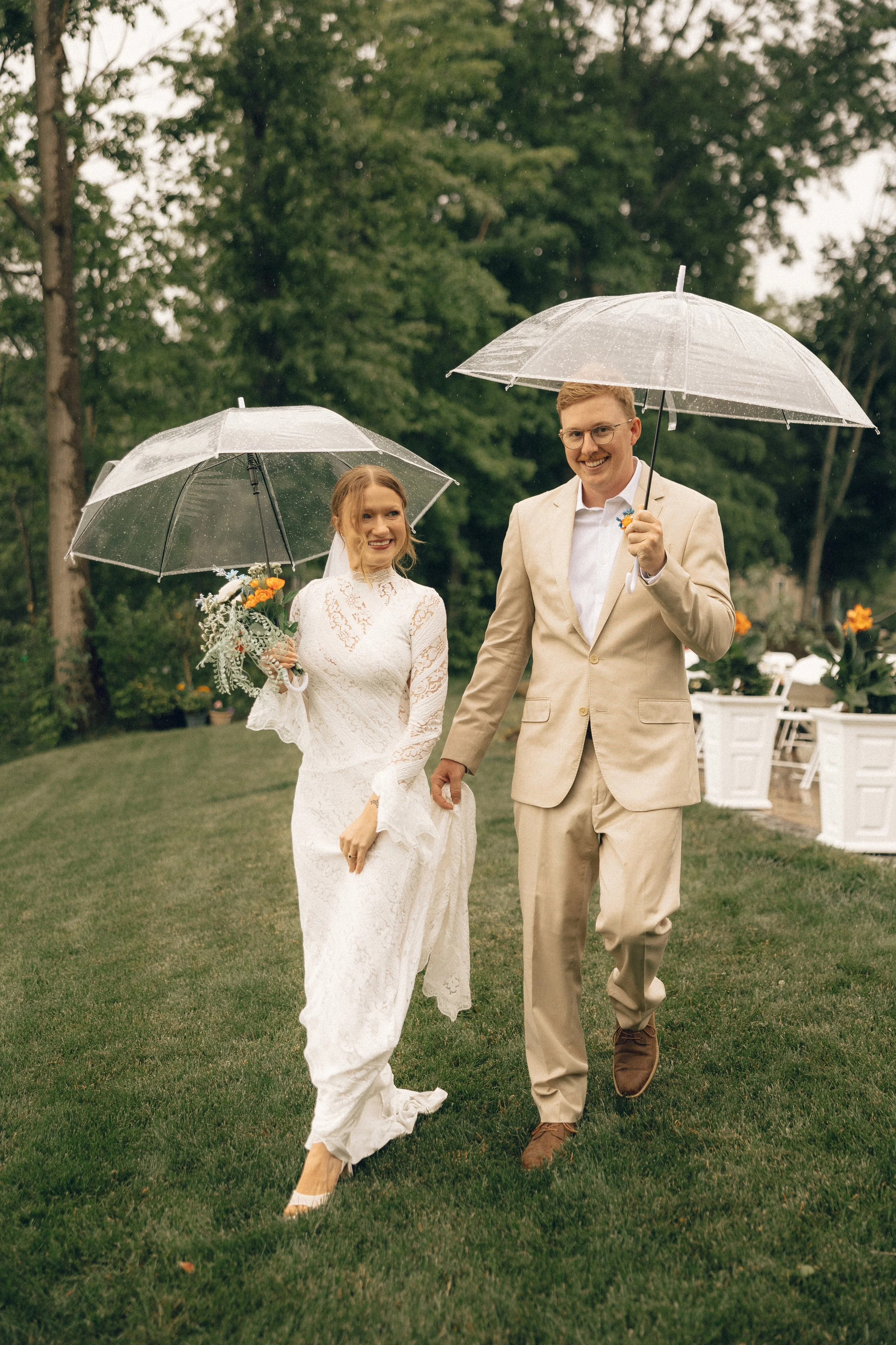 A bride and groom walking on grass in the rain, holding umbrellas. The bride is in a white lace wedding dress, holding a bouquet, and the groom is in a beige suit with glasses, holding an umbrella. They are smiling and hand in hand, with trees and ou