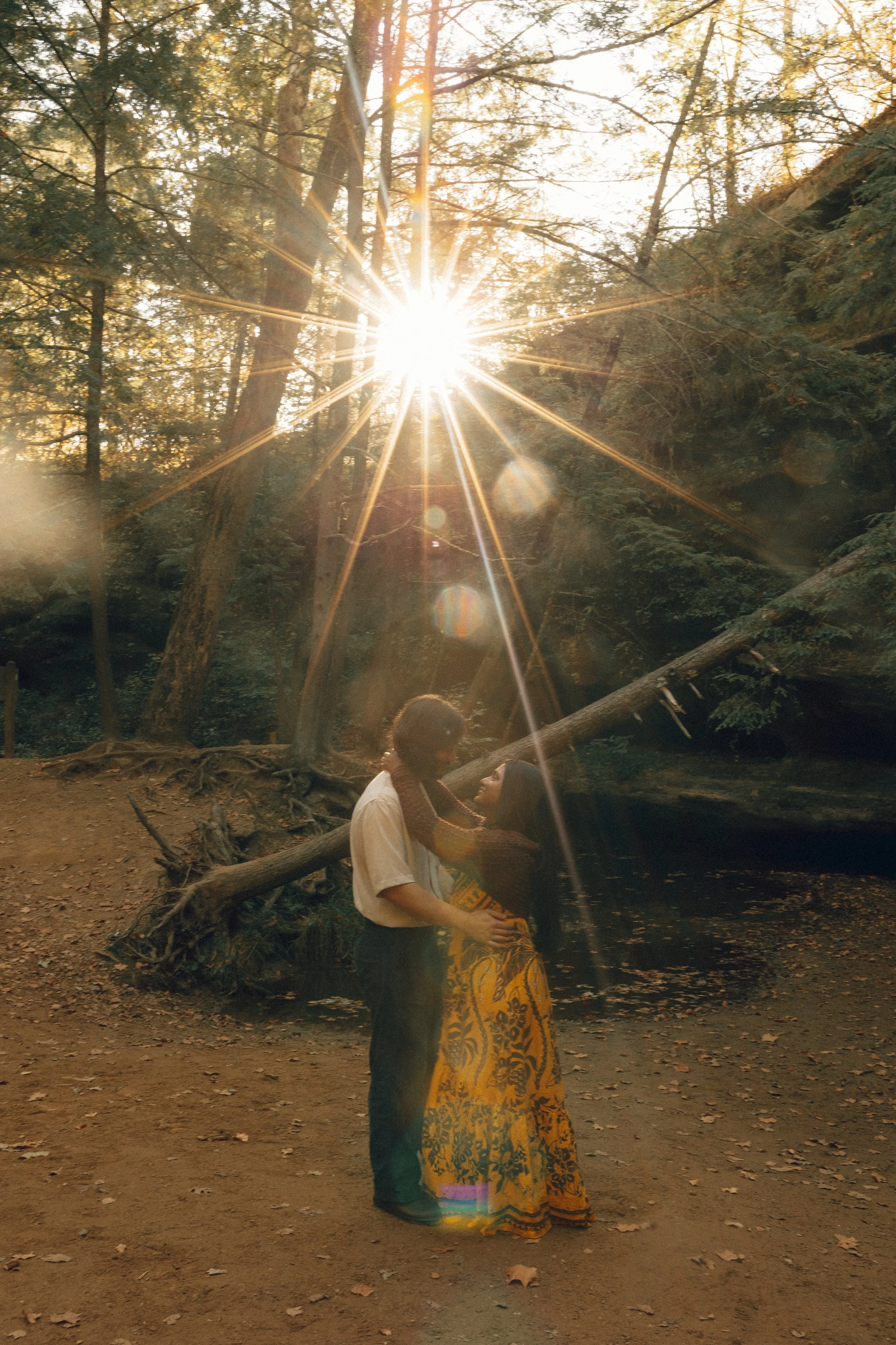 A couple stands closely together in a forest during sunset, holding hands and looking into each other's eyes. Sunlight filters through the trees, creating a starburst effect.