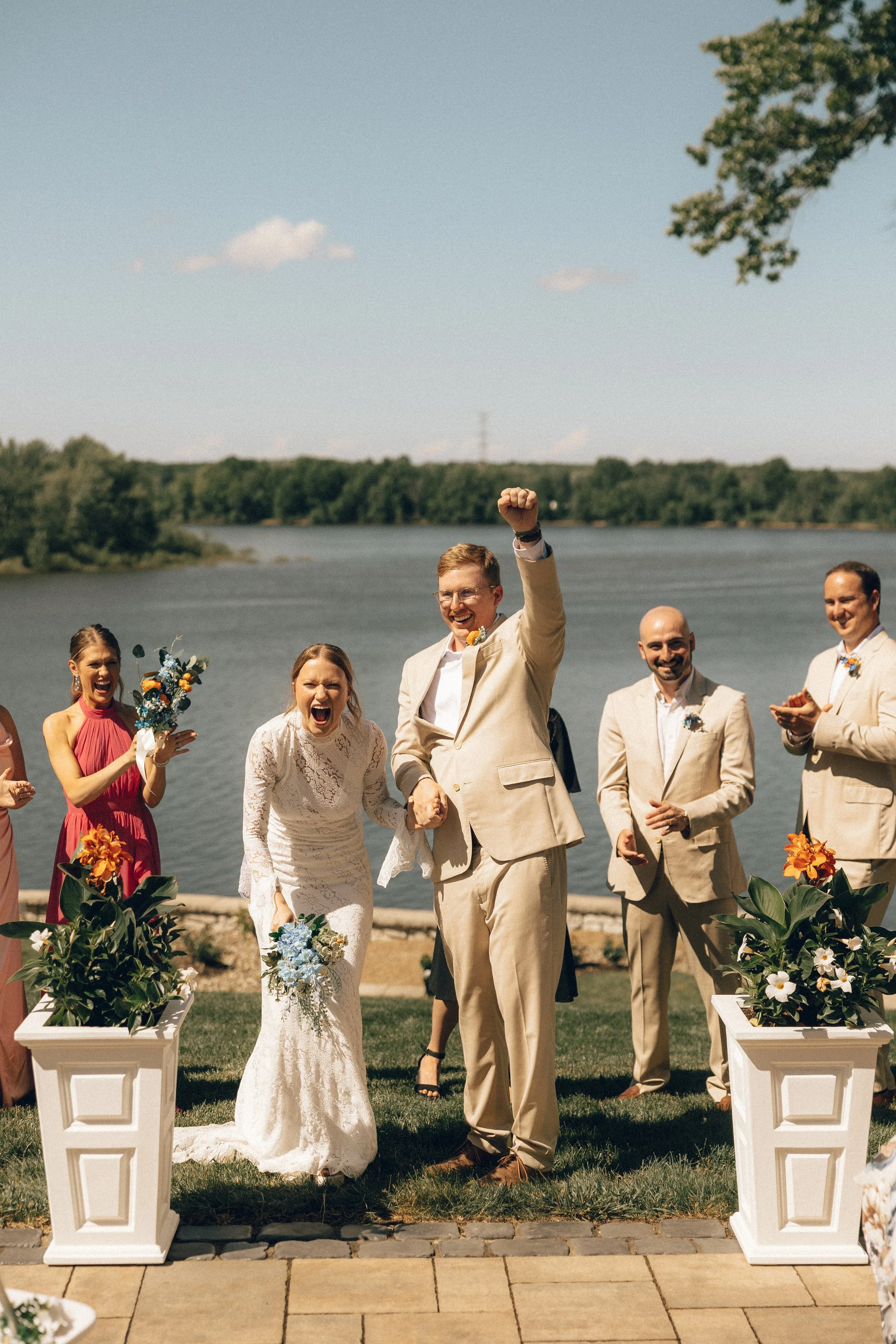 Bride and groom holding hands at an outdoor wedding ceremony by a lake, with bridesmaids and groomsmen smiling and standing behind them, decorated with flowers and greenery.