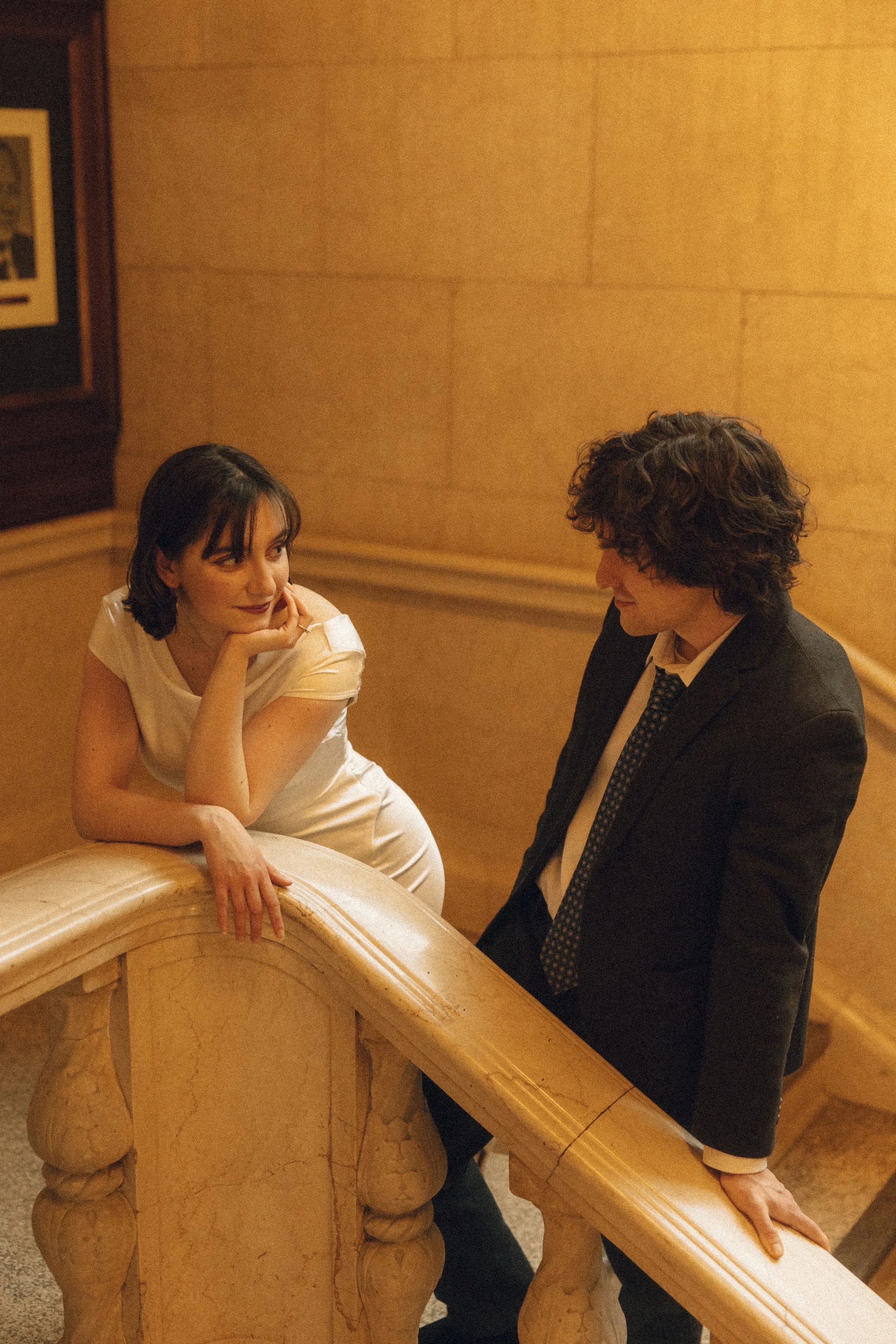 A woman sitting on a marble bench, resting her chin on her hand, and looking at a man standing beside her. The man is dressed in a dark suit and tie, and the woman is wearing a white dress. They are in a warmly lit indoor setting with wooden paneling