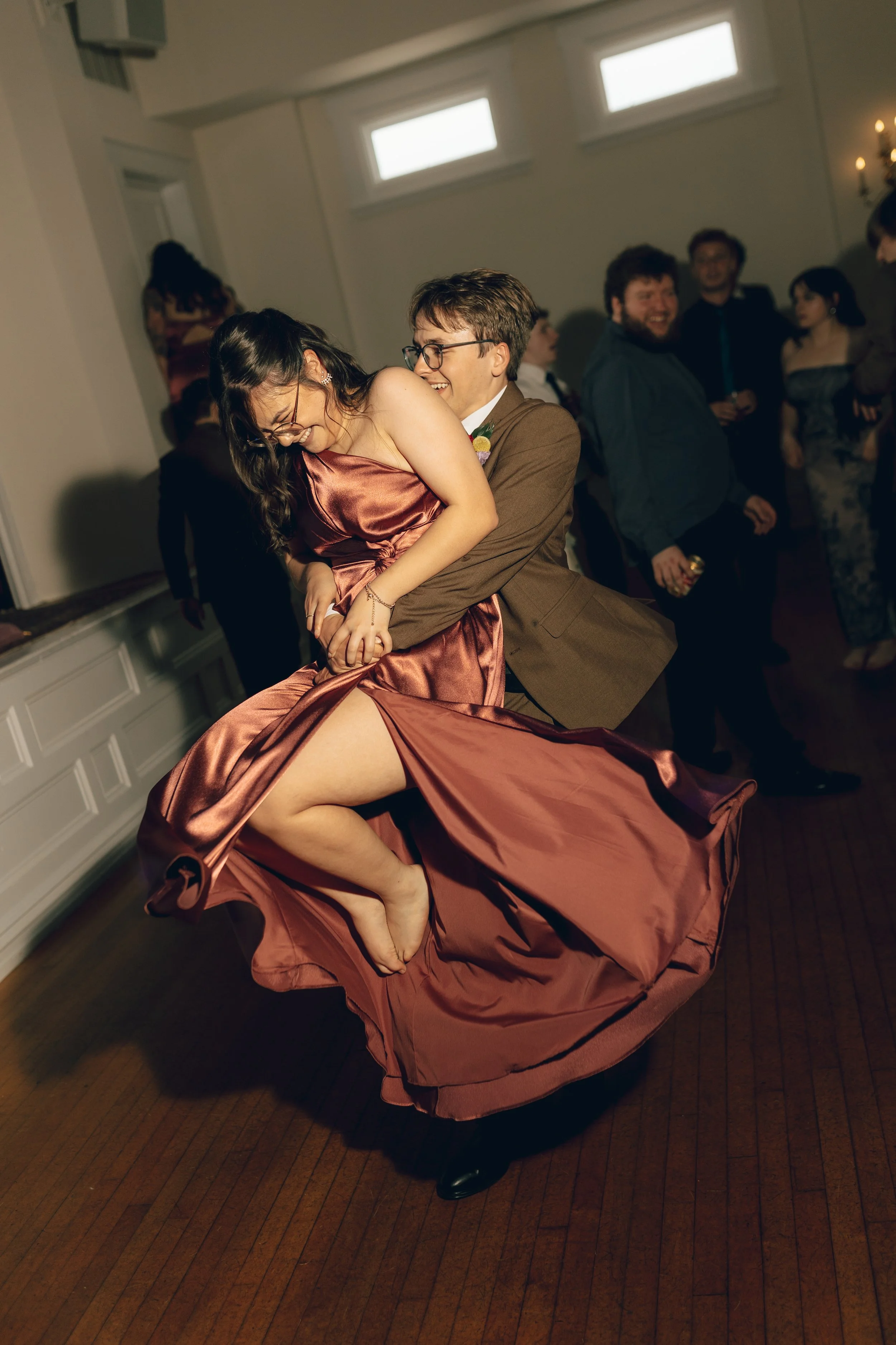 A woman in a satin dress dancing with a man wearing glasses and a suit at a party. Other guests can be seen in the background, smiling and socializing.