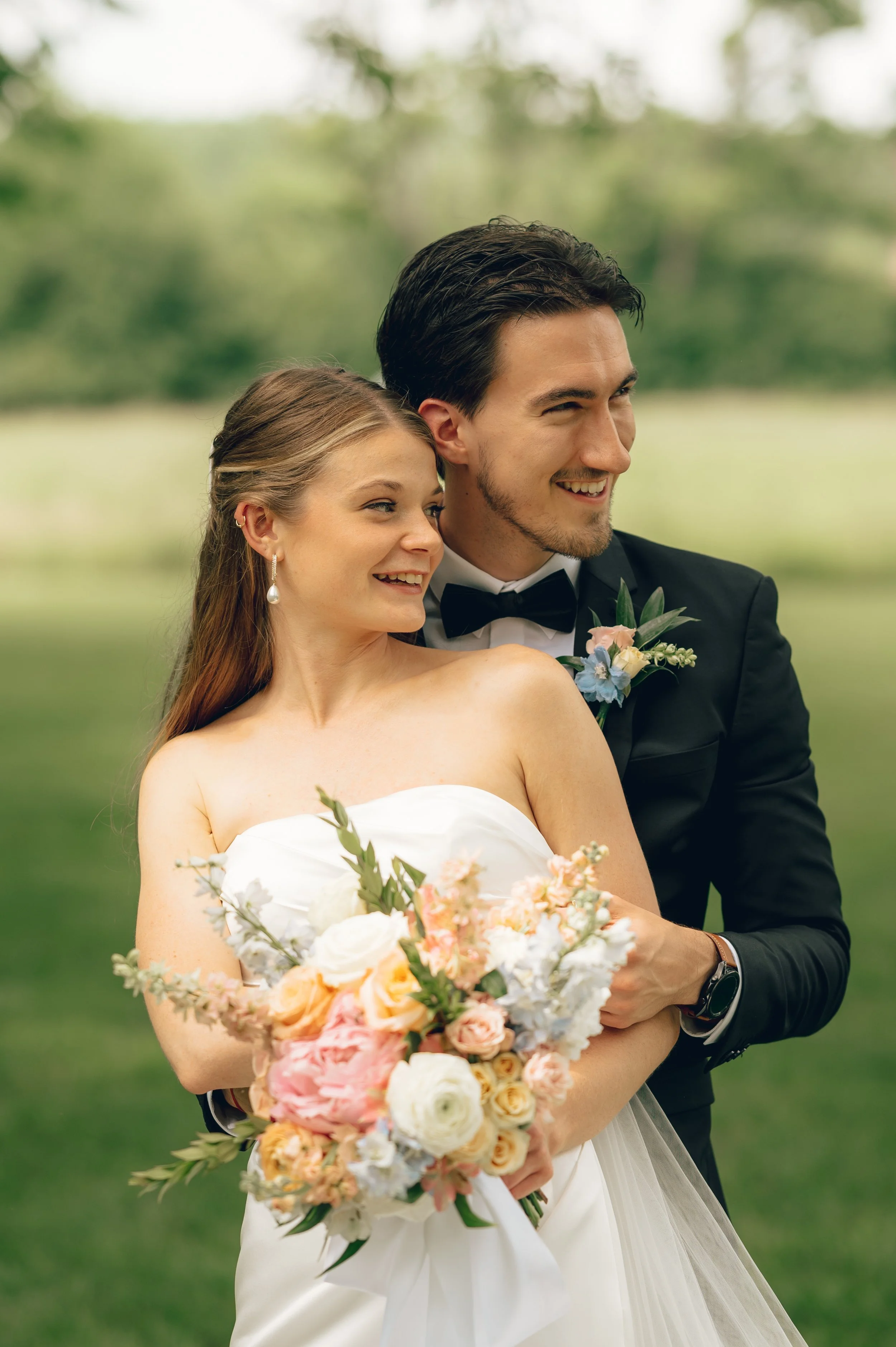 A bride and groom are embracing outdoors, smiling, with the bride holding a large bouquet of pastel-colored flowers, and the groom wearing a black tuxedo with a bow tie, in front of a blurred green background.